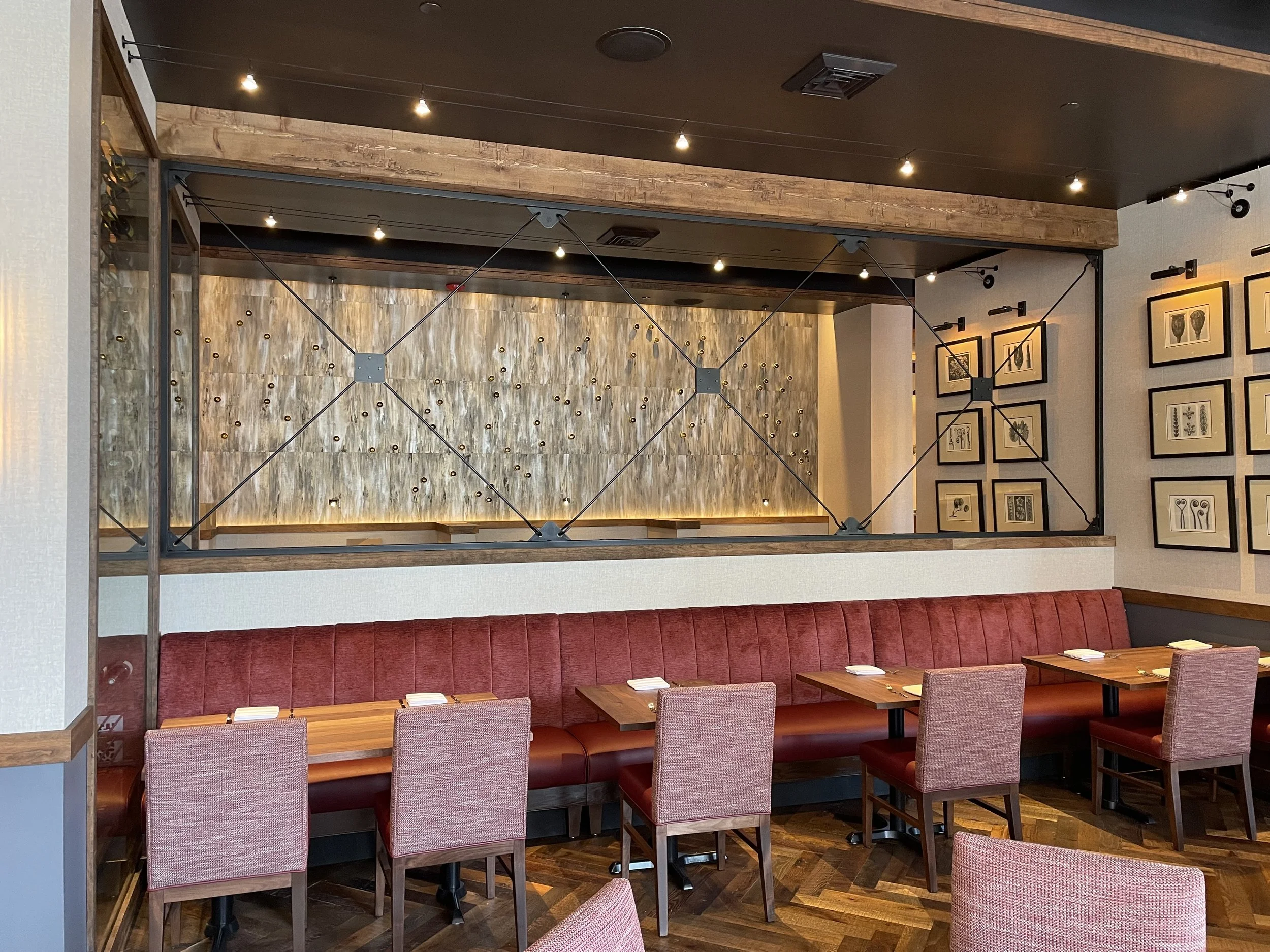 Empty restaurant with a long red upholstered bench along the wall, surrounded by individual pinkish chairs, wooden tables set with napkins and cutlery, framed artwork of plants and flowers on the wall, a decorative wooden partition, and small ceiling
