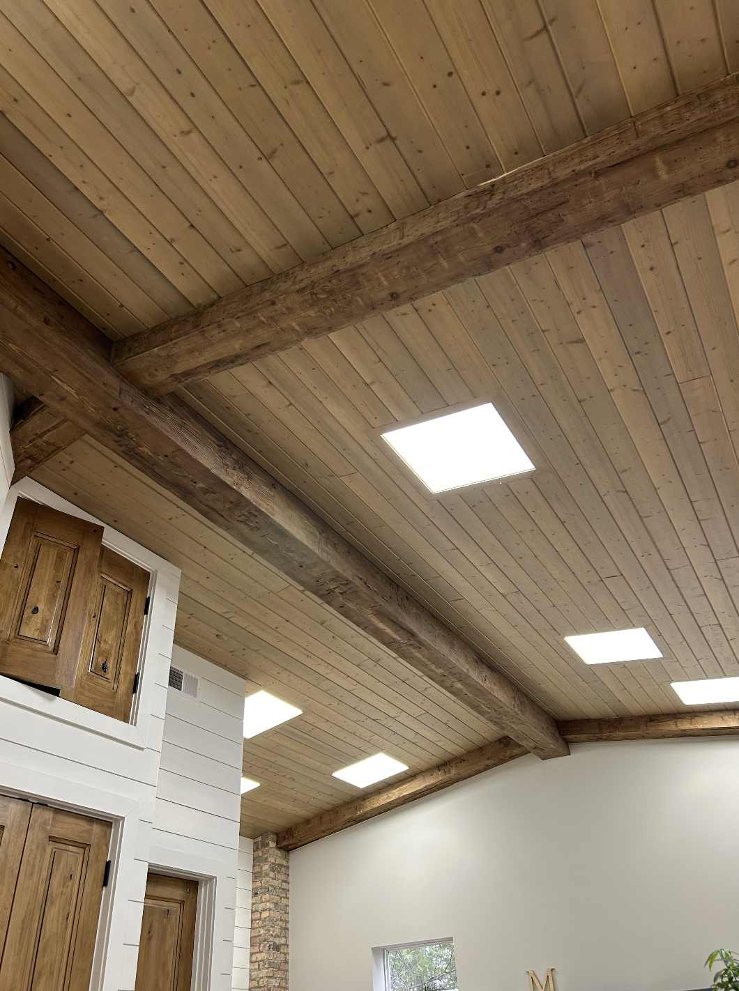 Interior of a room with wooden ceiling planks, exposed wooden beams, square ceiling lights, white wall, and wooden cabinet doors.