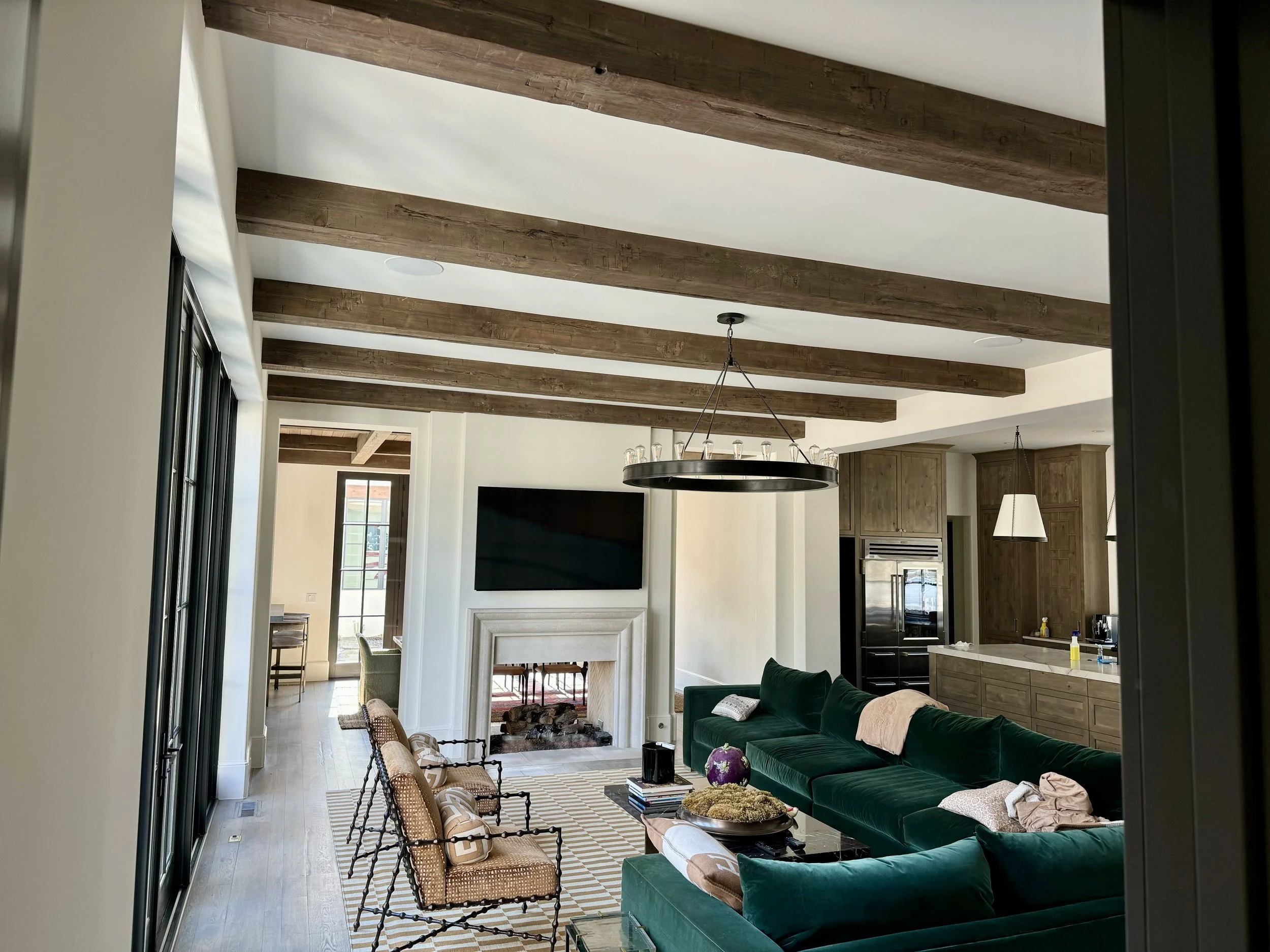 Living room with a dark green sectional sofa, a black coffee table, and beige armchairs with black frames. There is a white fireplace with a large flat-screen TV mounted above it, and a circular chandelier hangs from the wooden-beamed ceiling. The room has light-colored walls and hardwood floors.