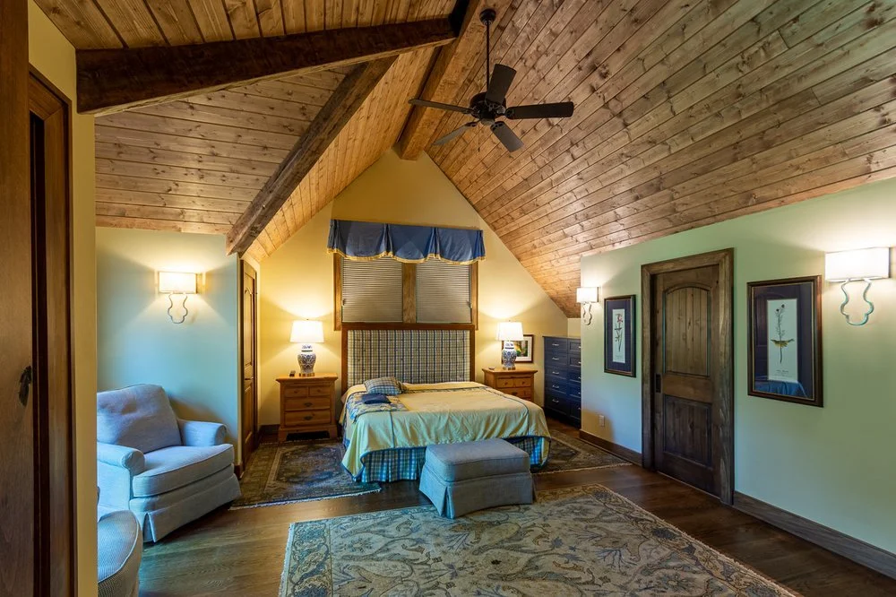 A cozy bedroom with a vaulted wooden ceiling, a ceiling fan, a bed with a checkered headboard, bedside tables with lamps, a sitting armchair, an ottoman, and framed artwork on the walls.