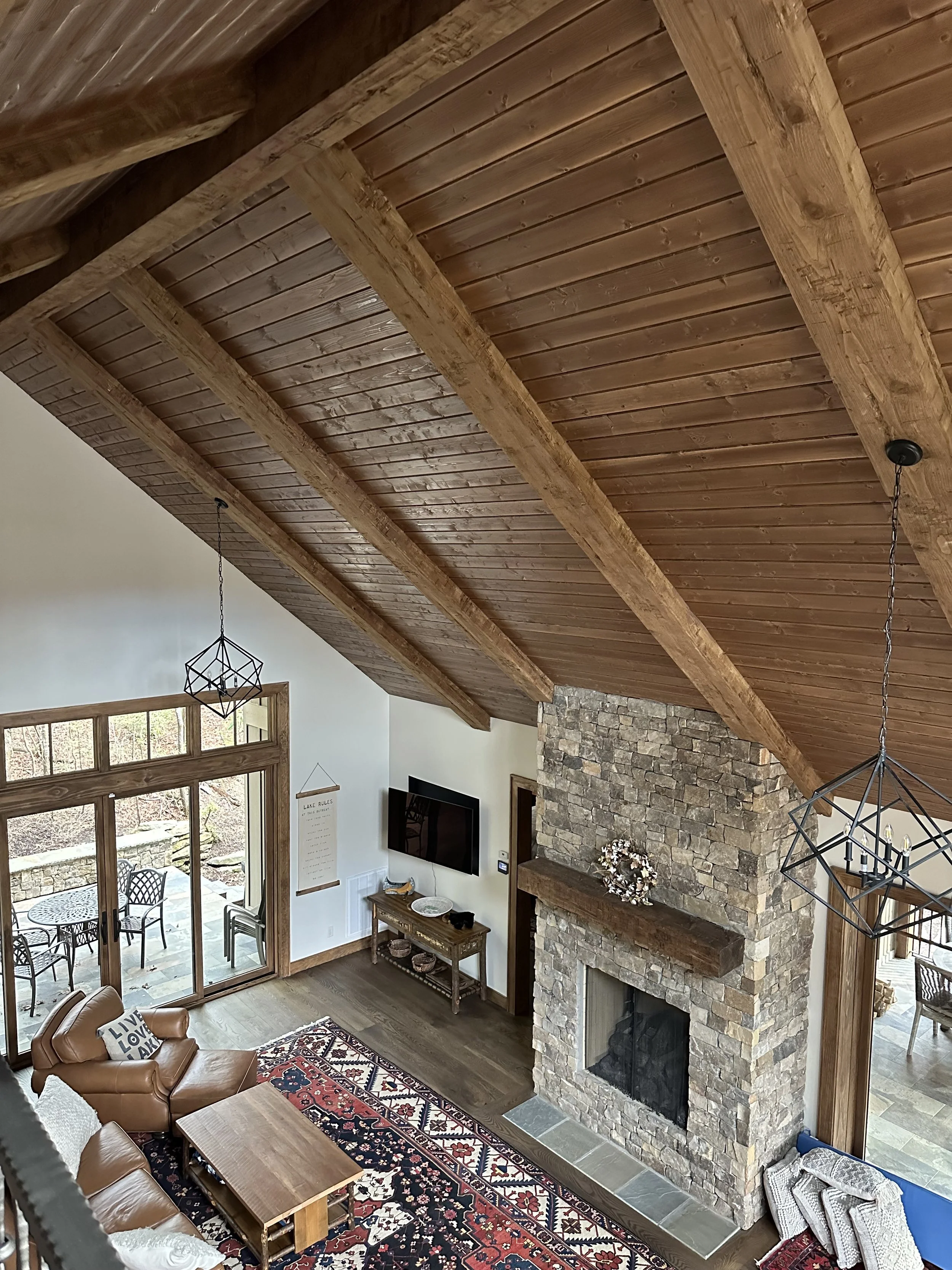Living room with a high wooden ceiling, stone fireplace, wall-mounted TV, glass sliding door leading to a patio, and seating area with a leather couch and patterned rug.