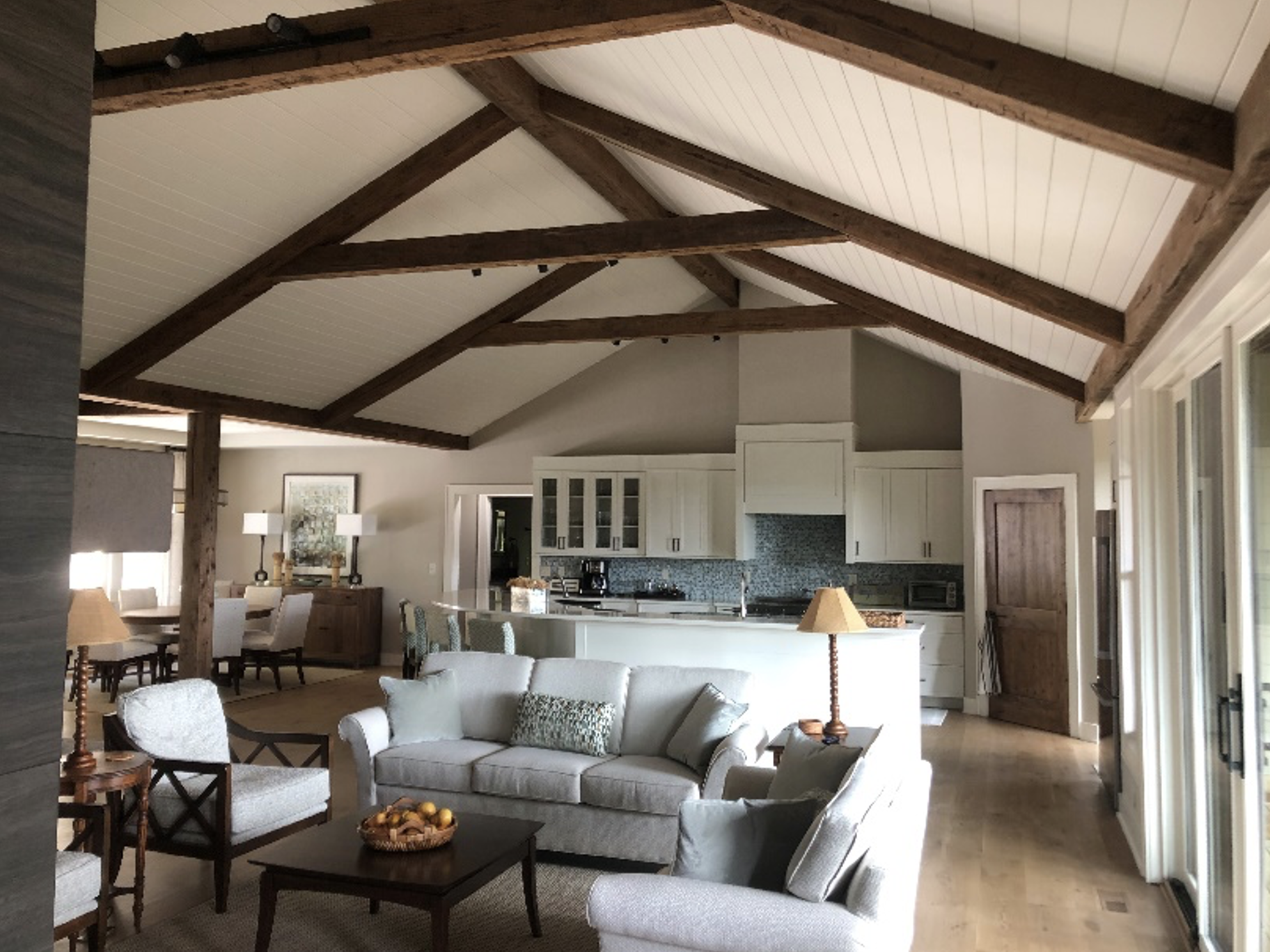 Living room with white sofa, armchair, and wooden side table with a lamp. Open kitchen with white cabinets, granite countertops, and a tiled backsplash. Wooden beams on the vaulted ceiling, sliding glass doors to the outside, and a dining area in the
