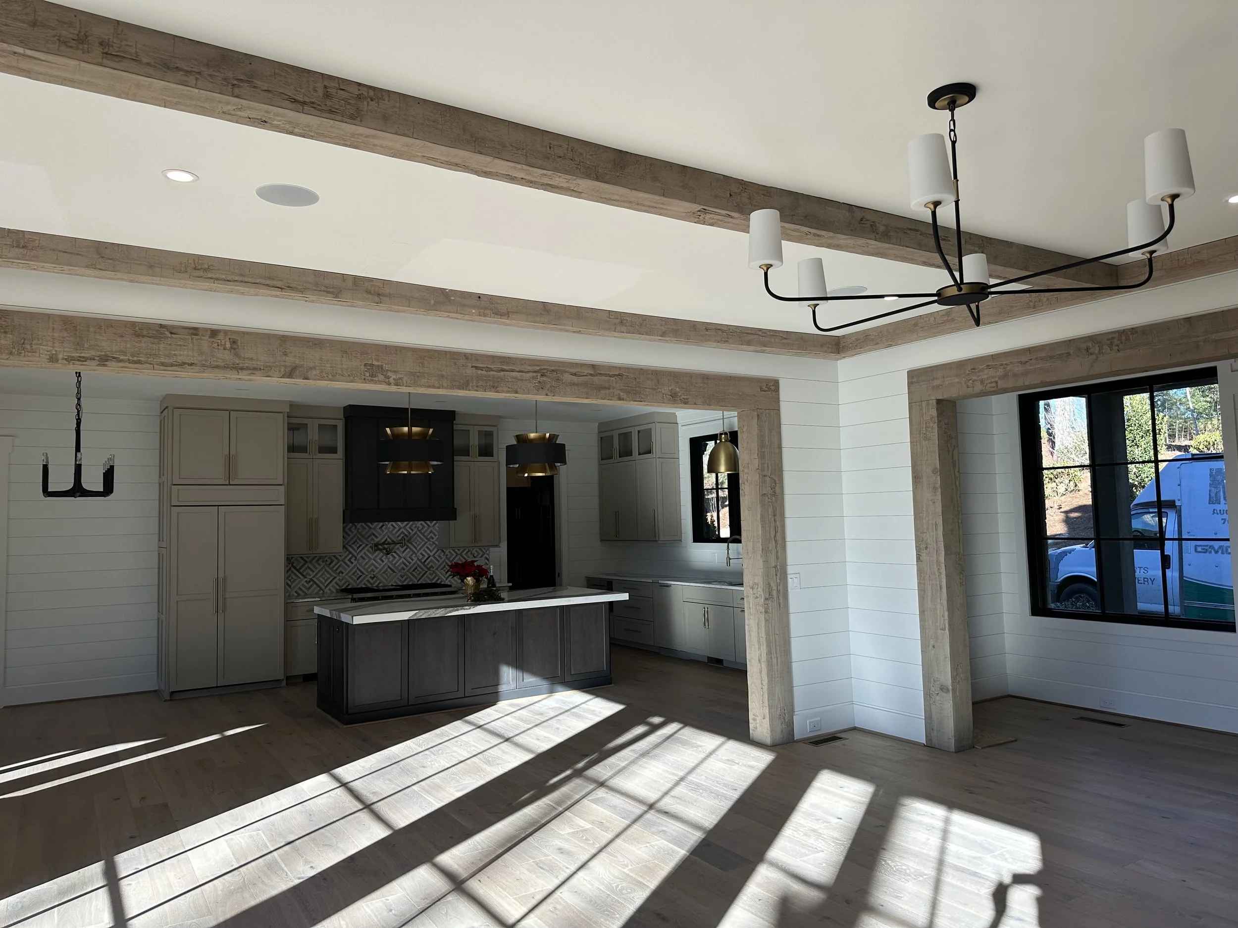 Open-concept kitchen and dining area with large window, wooden beams on ceiling, modern chandelier, and shadowed hardwood floors.