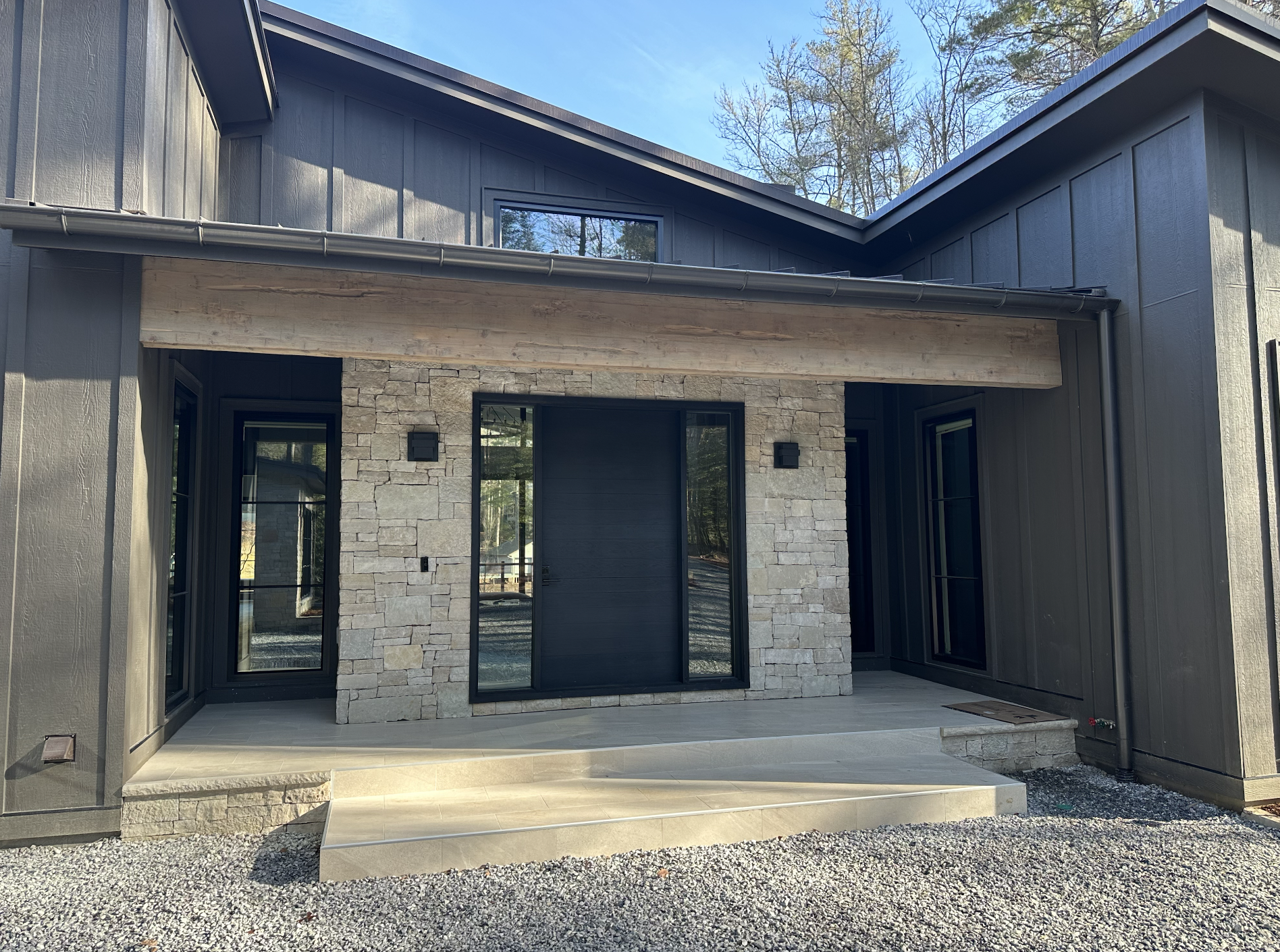 Modern house exterior with dark siding, stone accents, large black-framed glass doors, and small steps leading to patio, surrounded by gravel with trees in the background.
