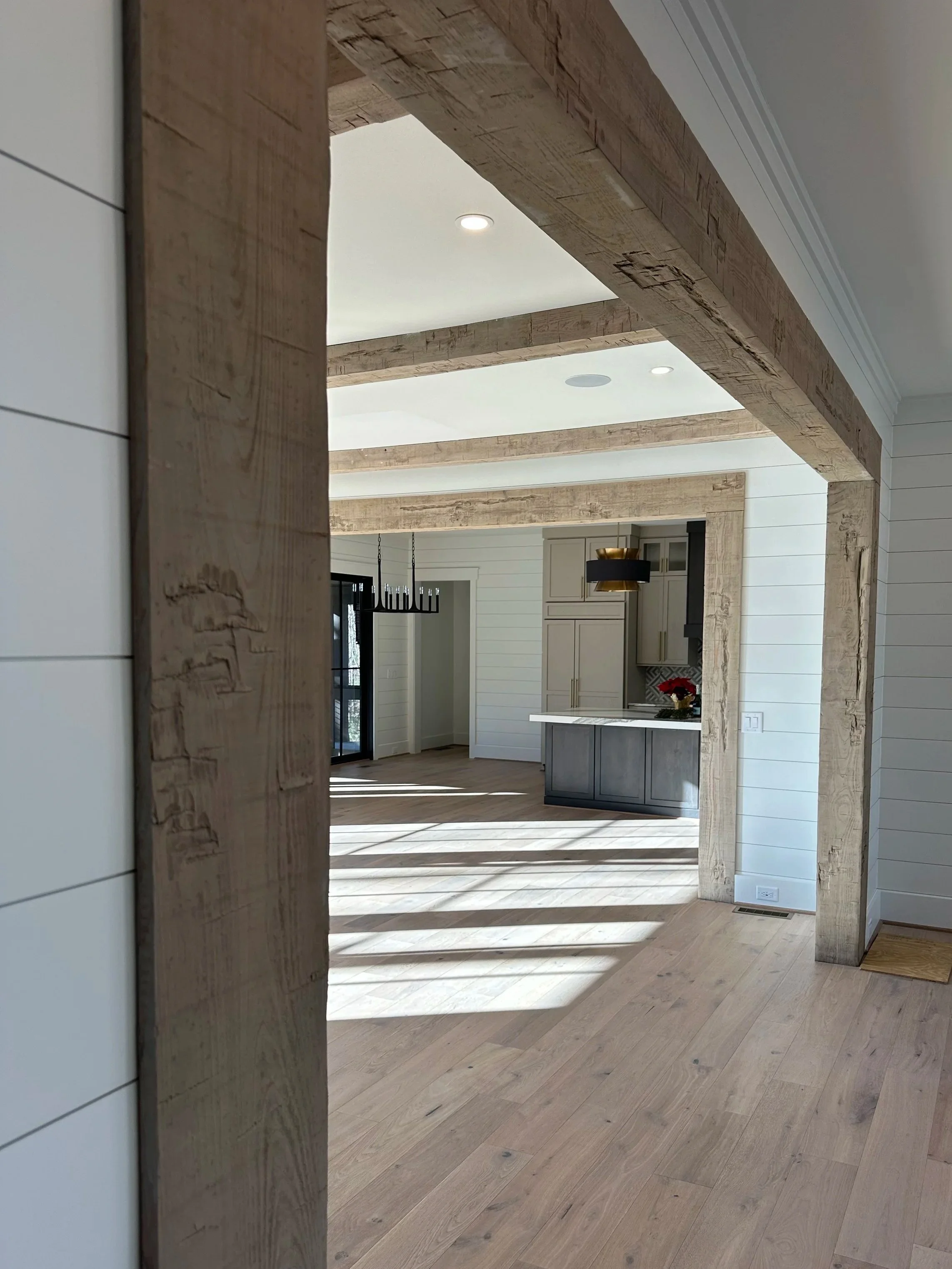 Open view of a modern kitchen and living space with wooden beams, white shiplap walls, and sunlight streaming through large windows and glass doors.