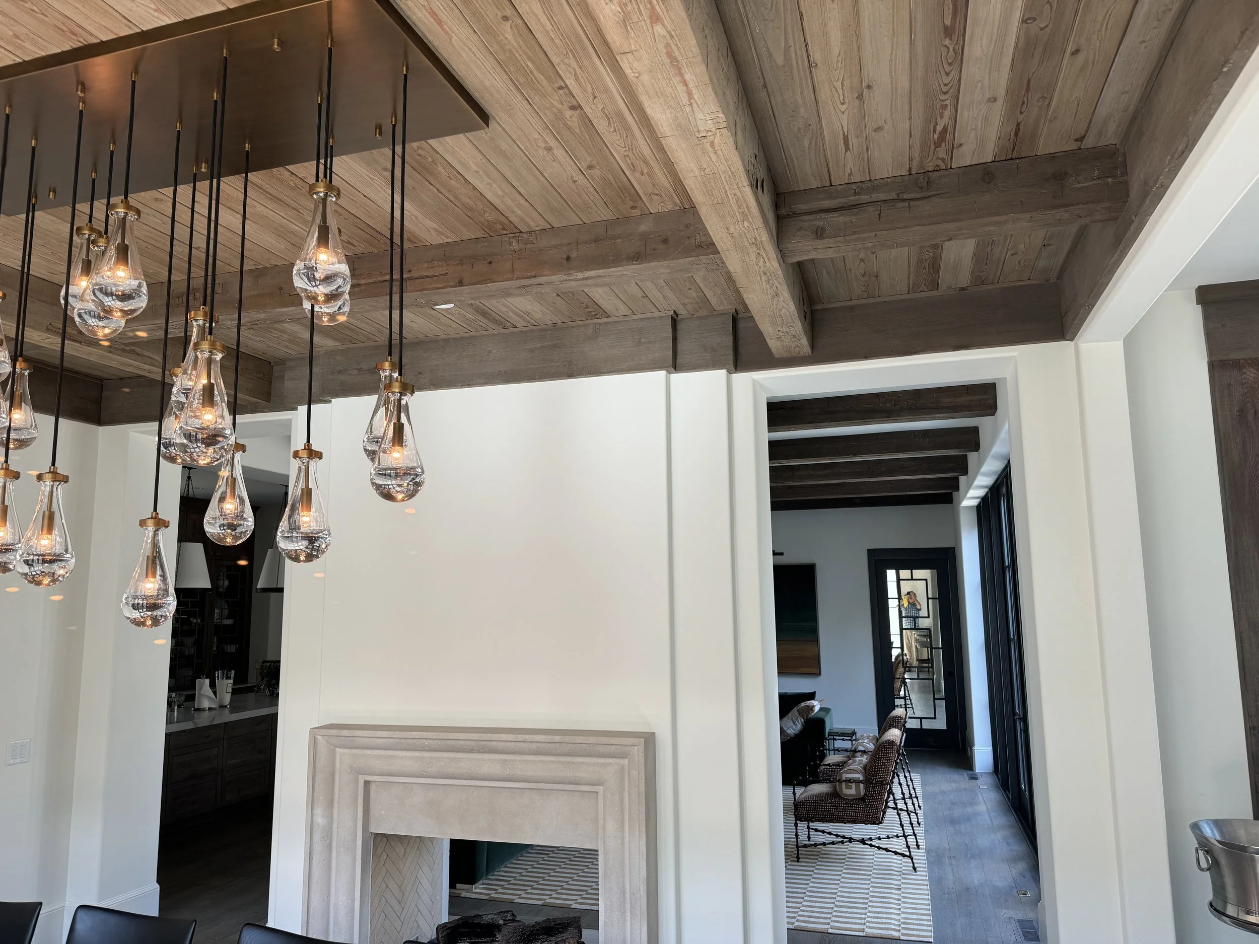 Open-concept living room with wooden ceiling beams, modern pendant lights, and a view into a sitting area with armchairs and a side table.