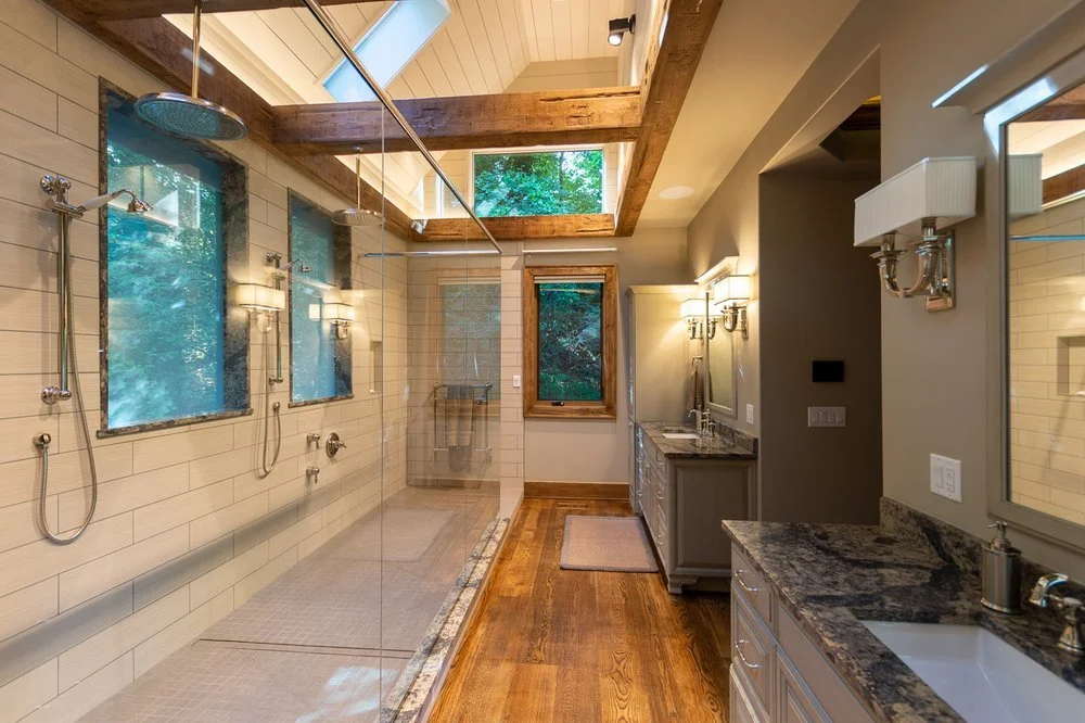 Bathroom with a spacious walk-in shower featuring three showerheads and windows showing trees outside. The bathroom also has a granite countertop sink, decorative wall sconces, mirrors, and wooden accents.
