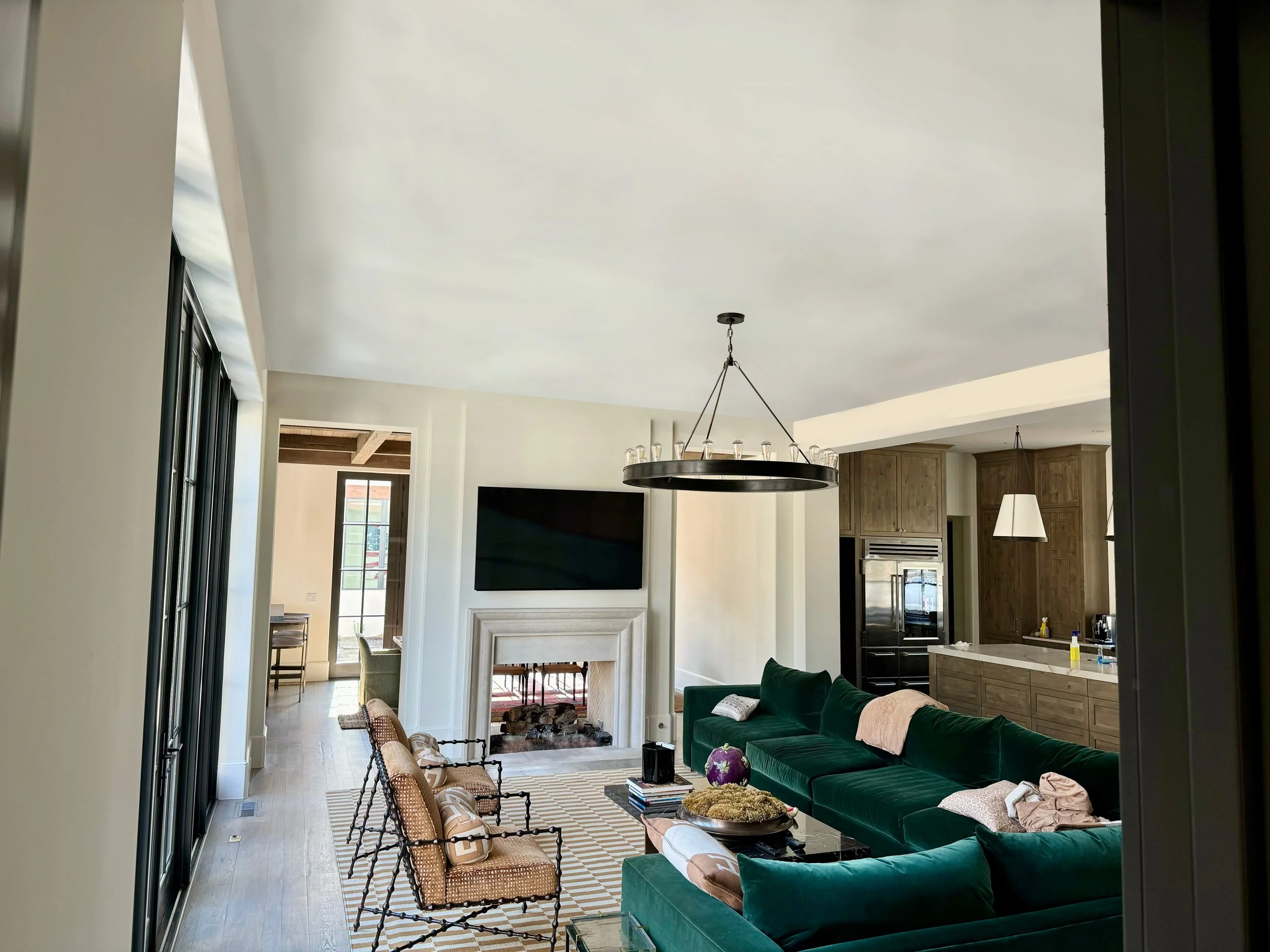 Living room with green velvet sectional, wooden chairs, a fireplace, a large wall-mounted TV, and a modern chandelier. Open kitchen area with wooden cabinets and black appliances visible in the background.