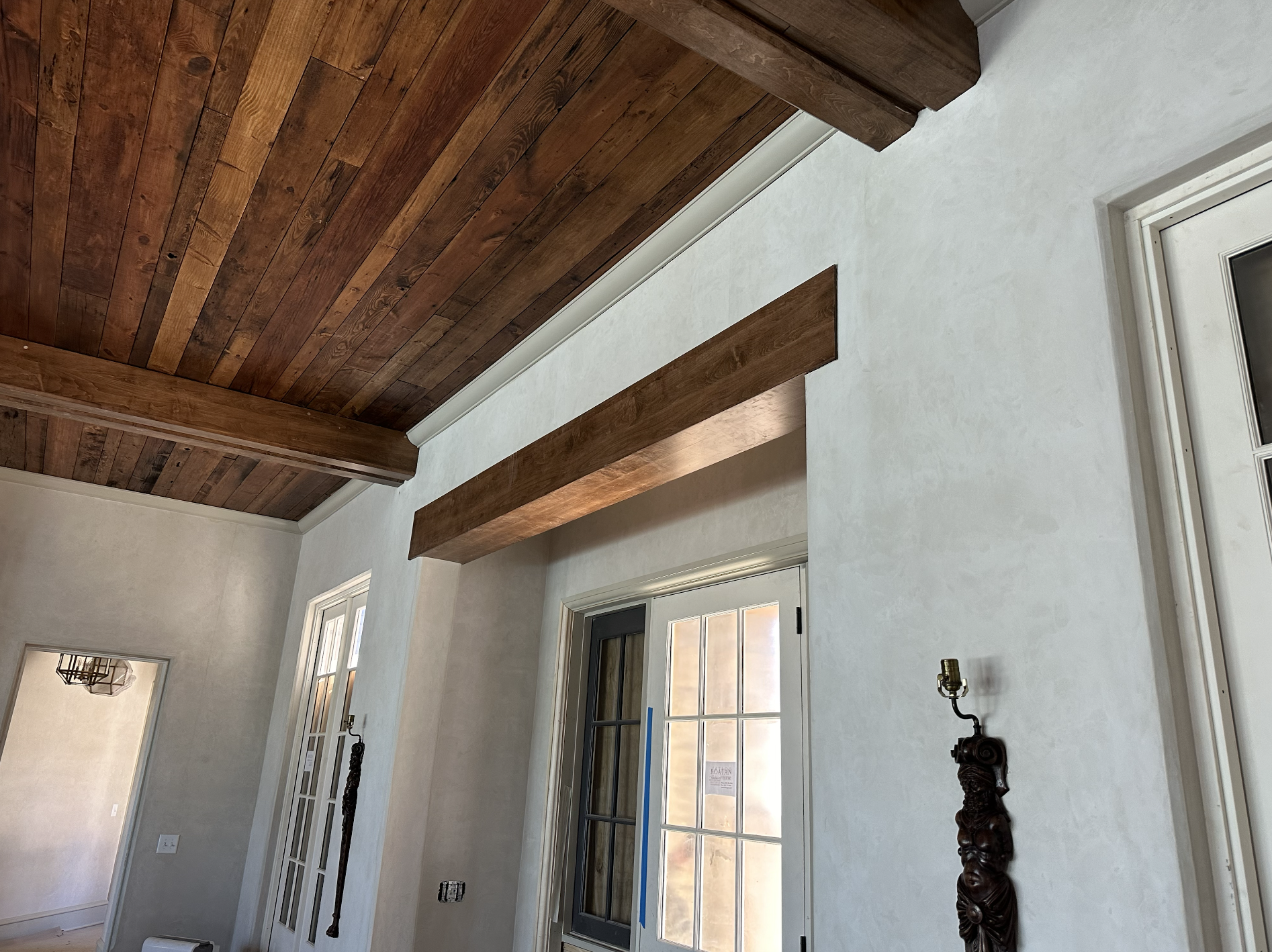 Interior view of a room with a wooden ceiling, white textured walls, a window, a door with glass panes, and decorative wooden statues.