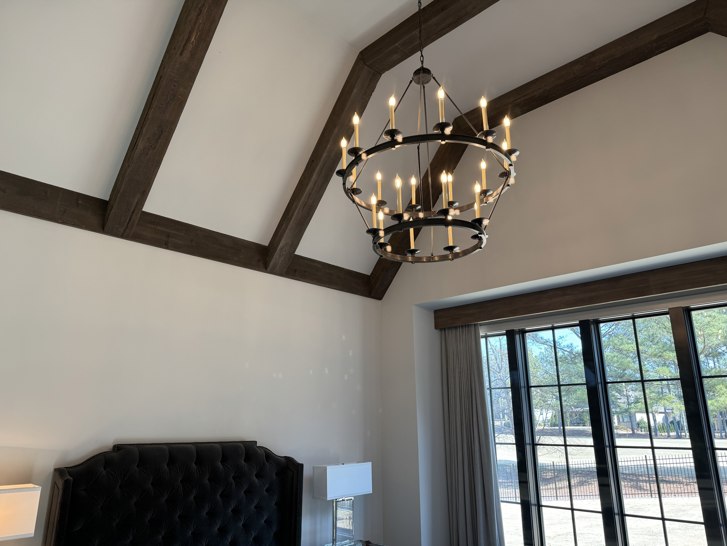 Interior of a room with vaulted ceiling, wooden beams, a black chandelier with multiple candle-shaped lights, a large window with curtains, and a bed with a tufted headboard.