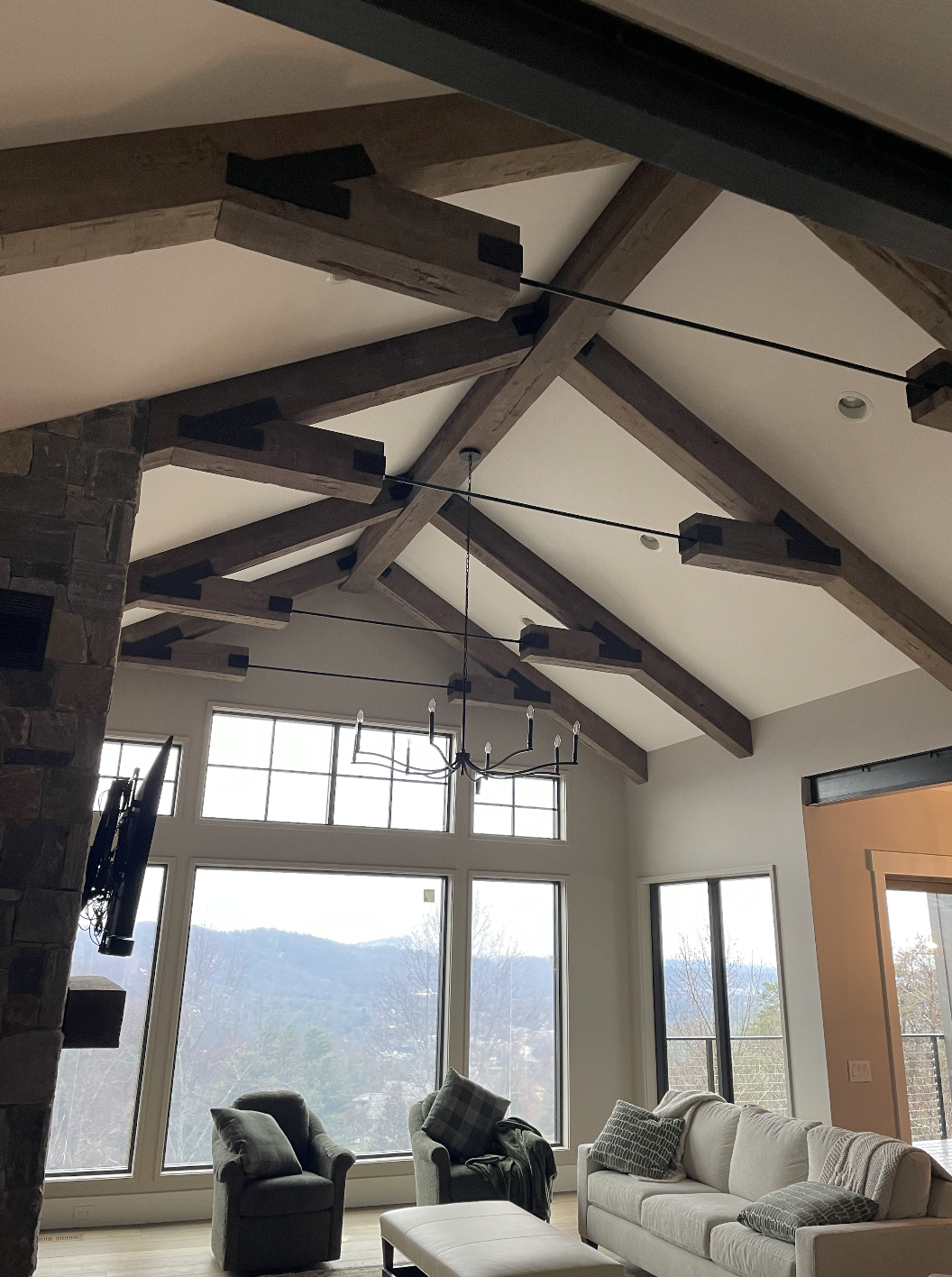 Living room with large window showing mountains, ceiling with wooden beams, chandelier, and sofa with pillows.