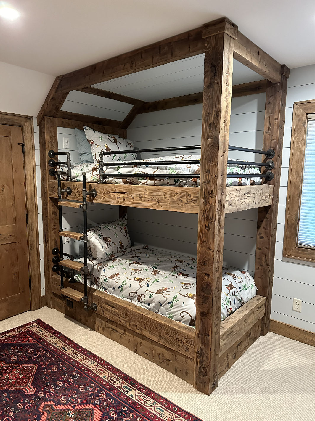 A wooden bunk bed with a rustic design in a room, featuring two beds with colorful monkey-themed bedding and a black metal ladder. The room has wooden doors, a window with wooden trim, and a patterned rug on the beige carpeted floor.