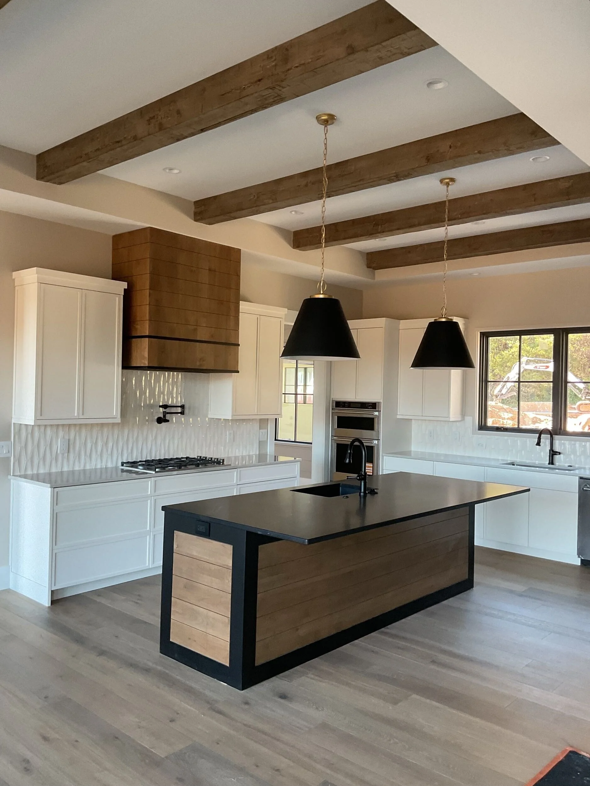 Modern kitchen with white cabinets, a black island with a wood panel accent, black pendant lights, wood beams on the ceiling, stainless steel appliances, and a window with a view outside.
