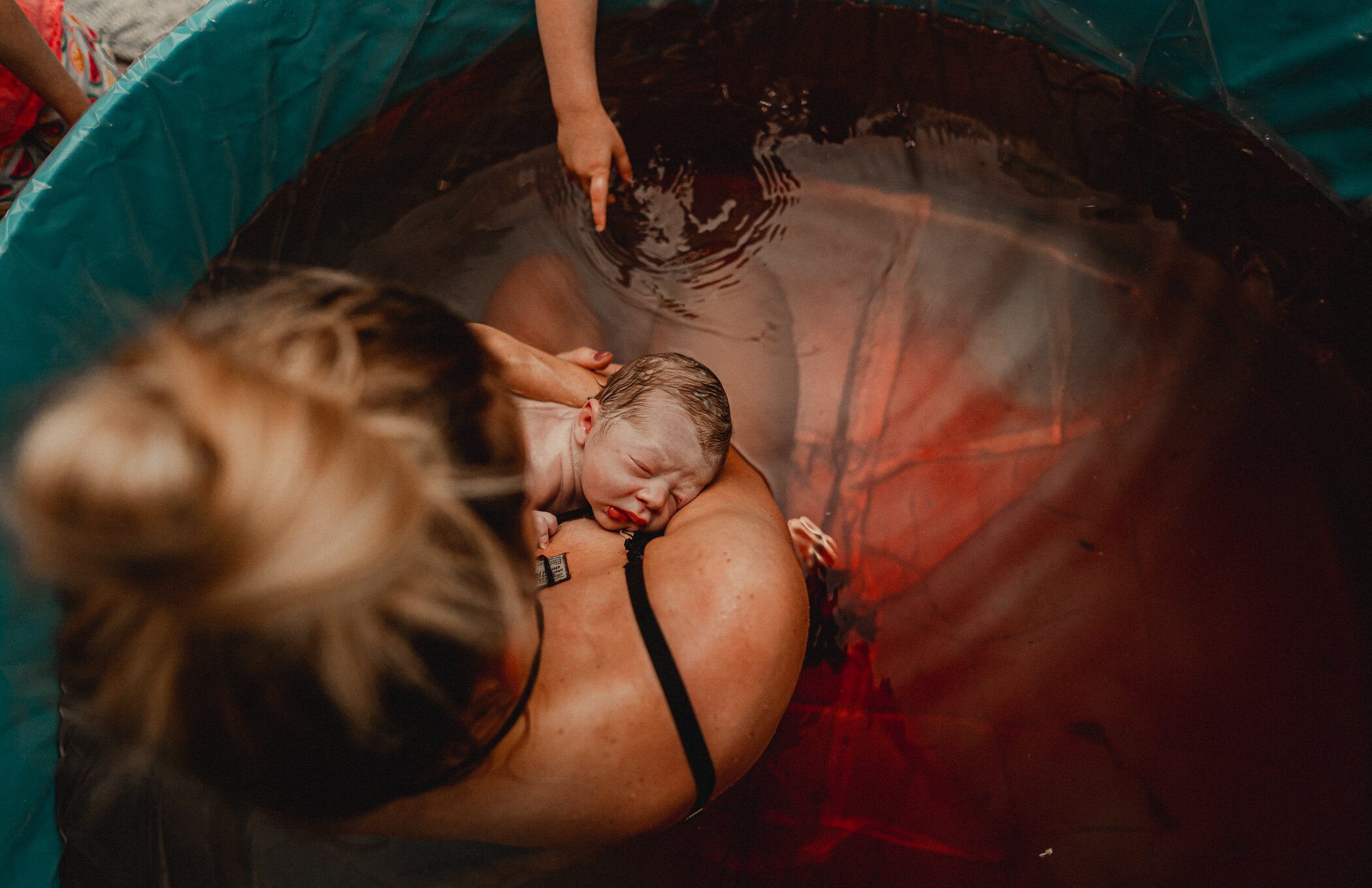 A woman is sitting in the birth pool holding her newborn son who is sleeping in her arms. A finger is reaching in eagerly waiting to touch baby brother
