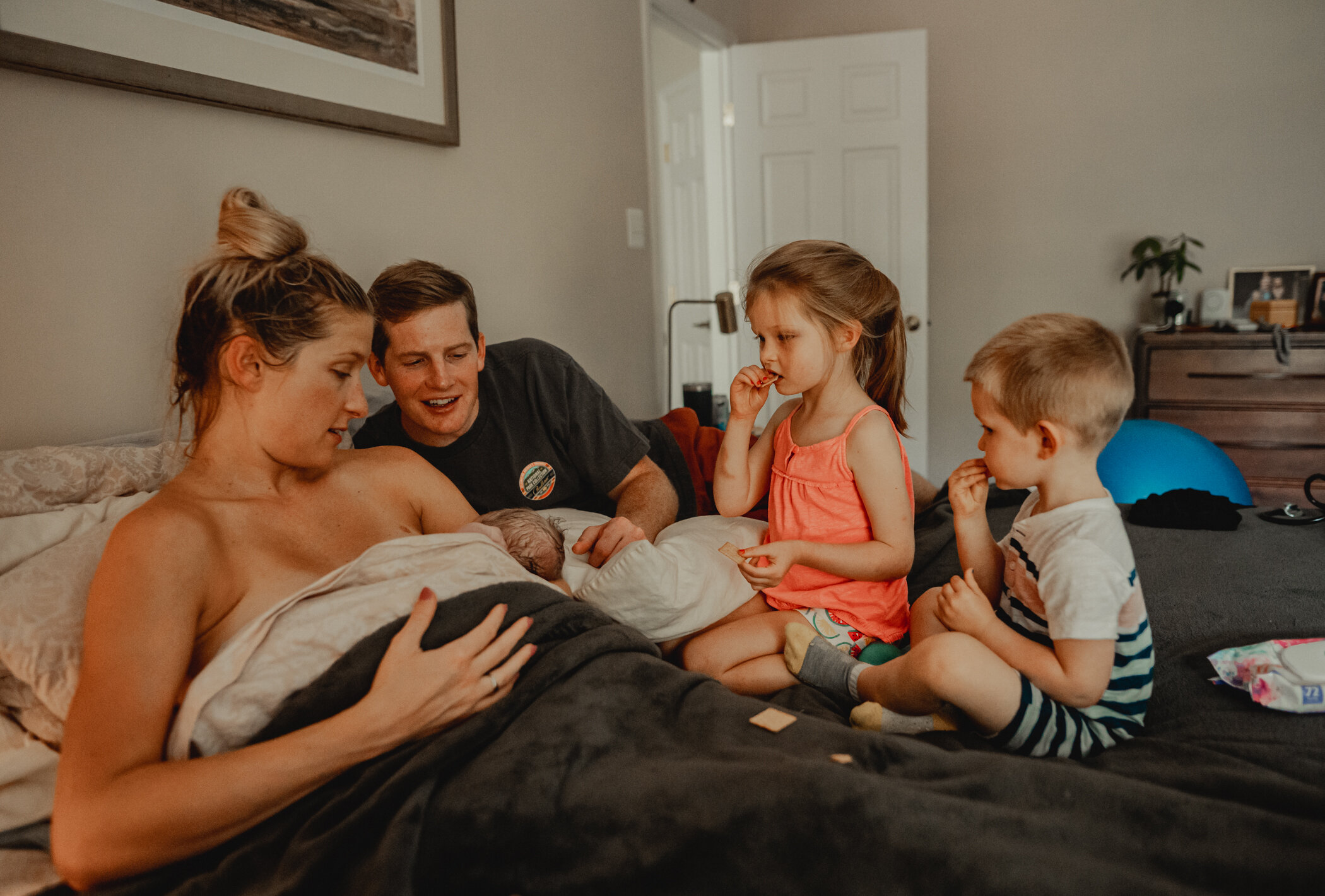 Family is sitting on the bed, older siblings are eating a snack and everybody is admiring the newest family addition who is nursing with mama after his fast home water birth in Great Falls, VA