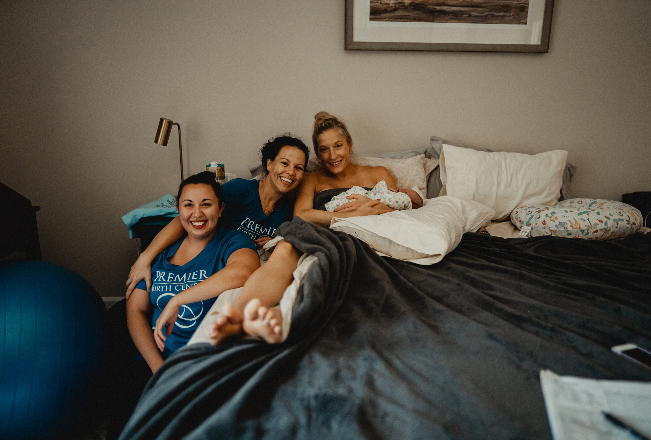 Mama is sitting in her bed with her sleeping new baby on her chest while her midwife,Mayanne and her birth assistant are sitting besides her. All women are looking at the camera 