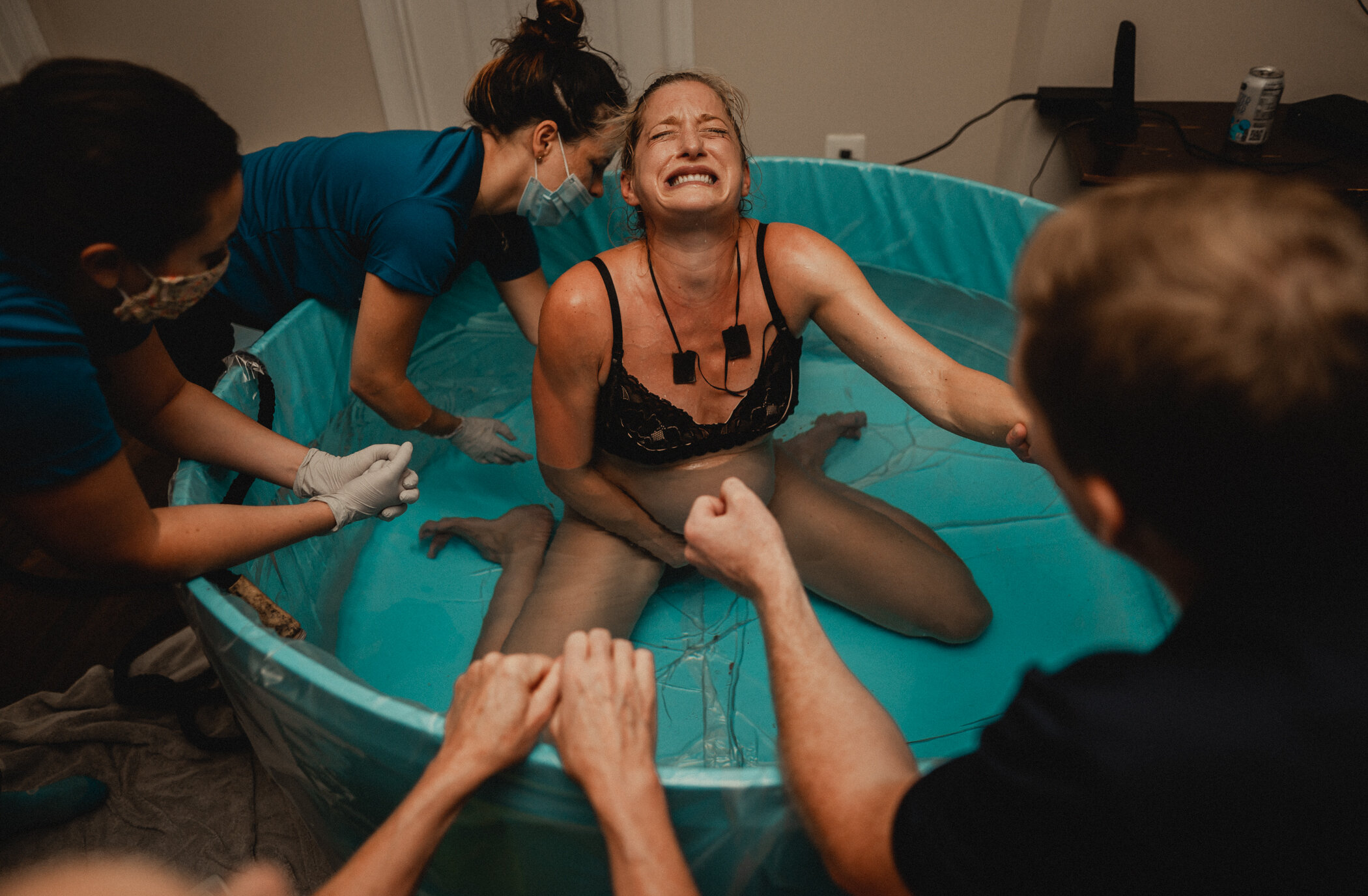 Woman sitting in the birth tub experiencing one last contraction before her baby's head is crowning. Her midwife kneeling behind her providing counter pressure