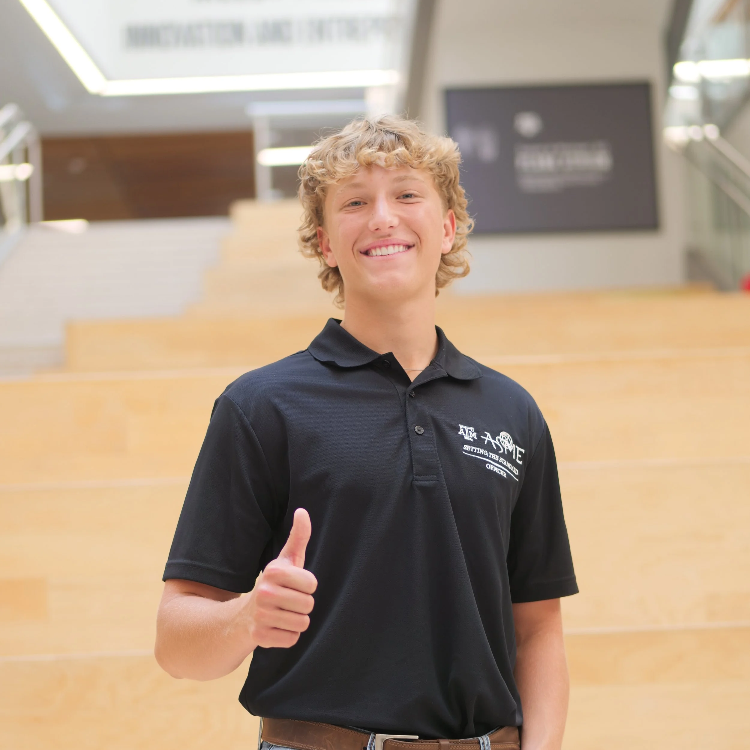 A smiling young man with curly blond hair giving a thumbs-up in an indoor setting with wooden stairs and a large black sign in the background.
