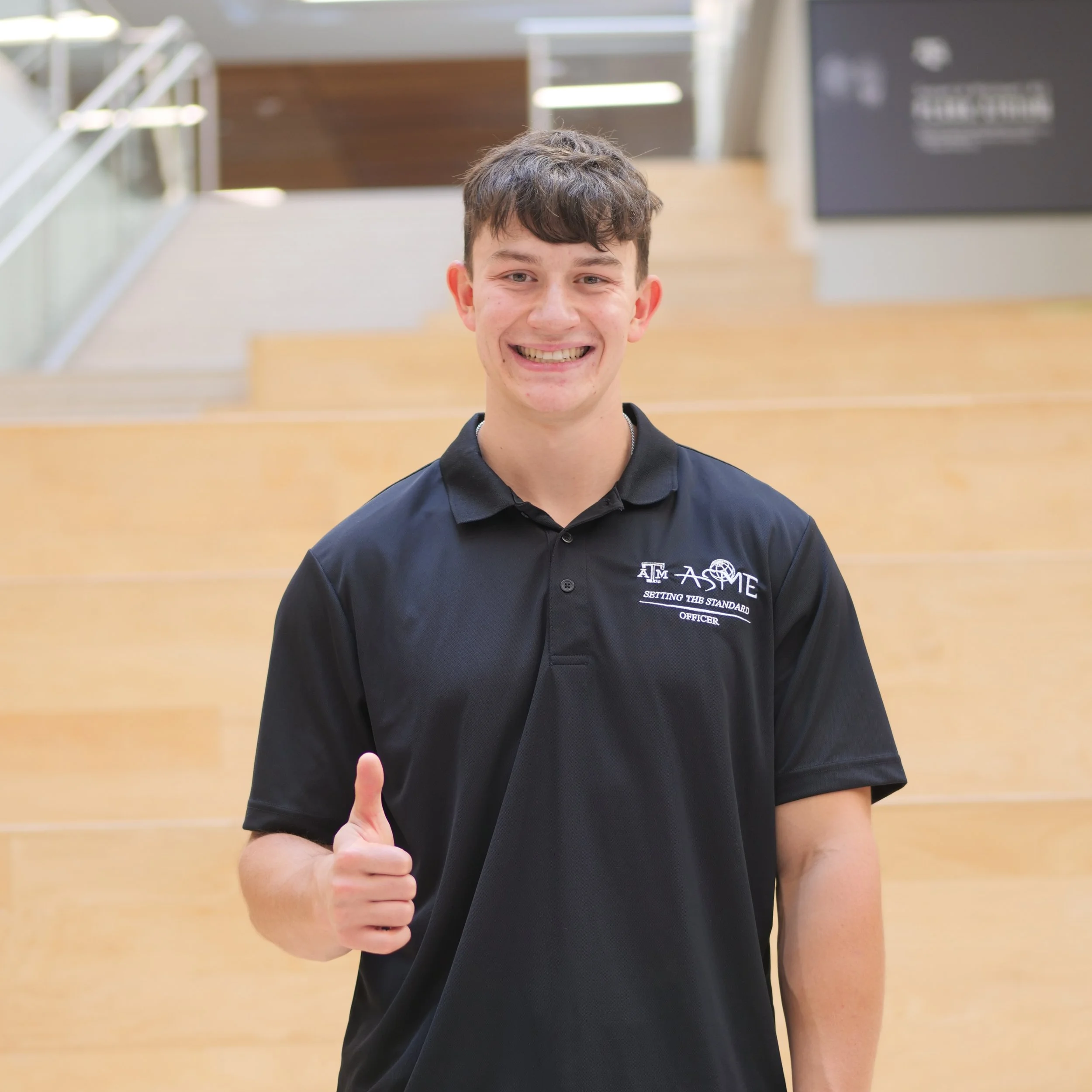 Young man smiling and giving a thumbs-up in an indoor setting, wearing a black polo shirt with an emblem and text.