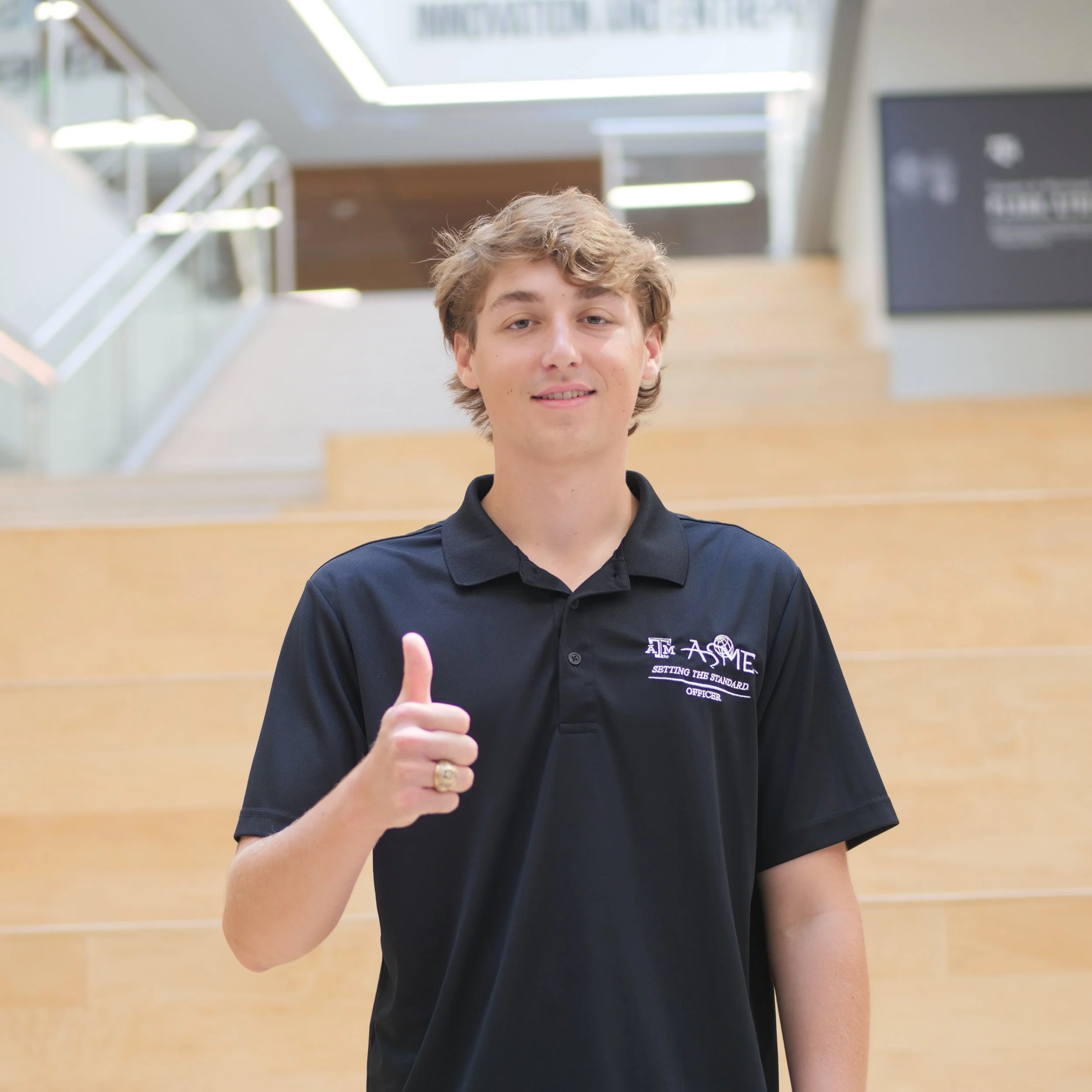 A young man in a black polo shirt with an emblem and text on it, giving a thumbs-up and smiling in a modern, well-lit building with stairs and glass railing.