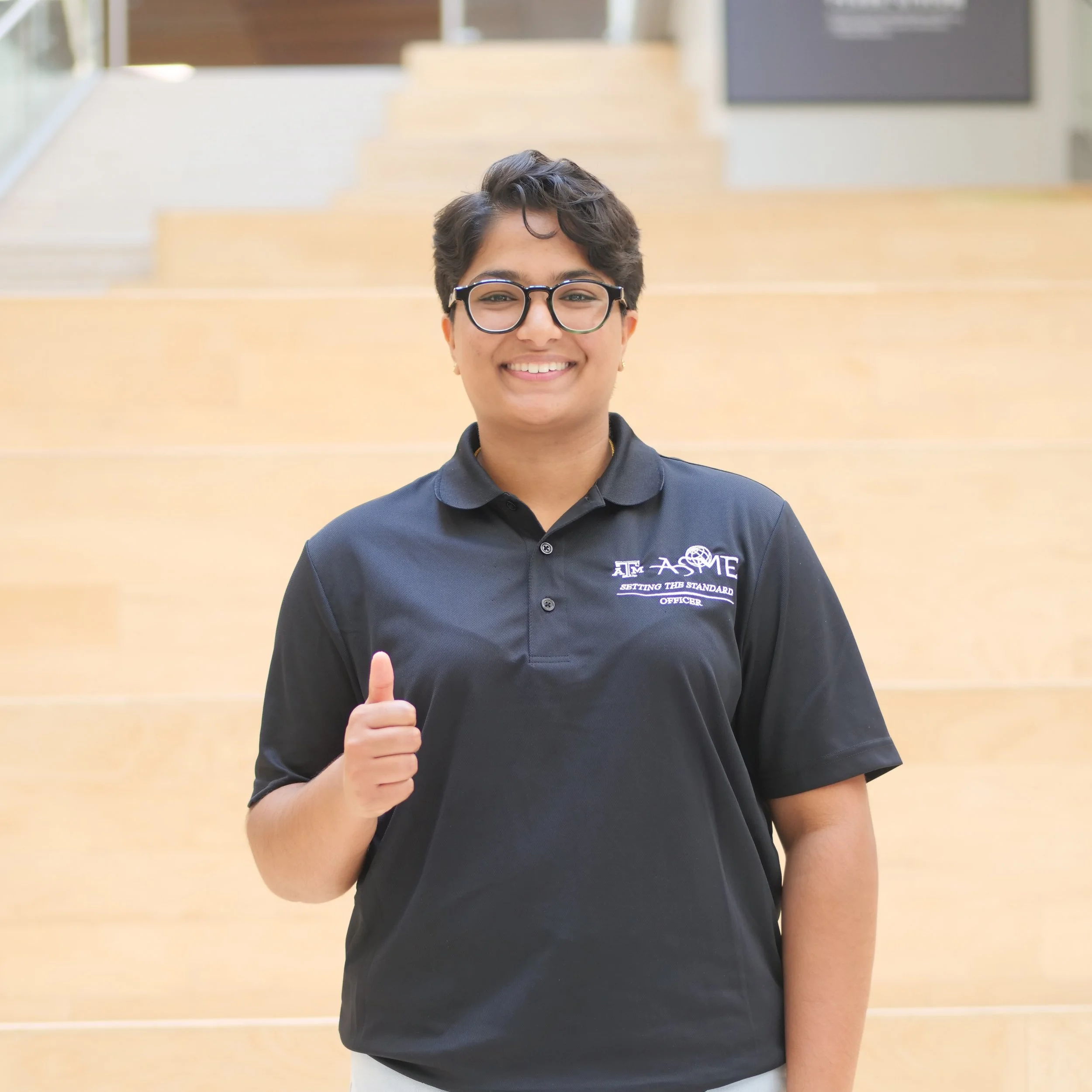 A young woman with short dark hair, glasses, and a black polo shirt with a logo and text, standing indoors in front of a staircase, smiling and giving a thumbs-up.