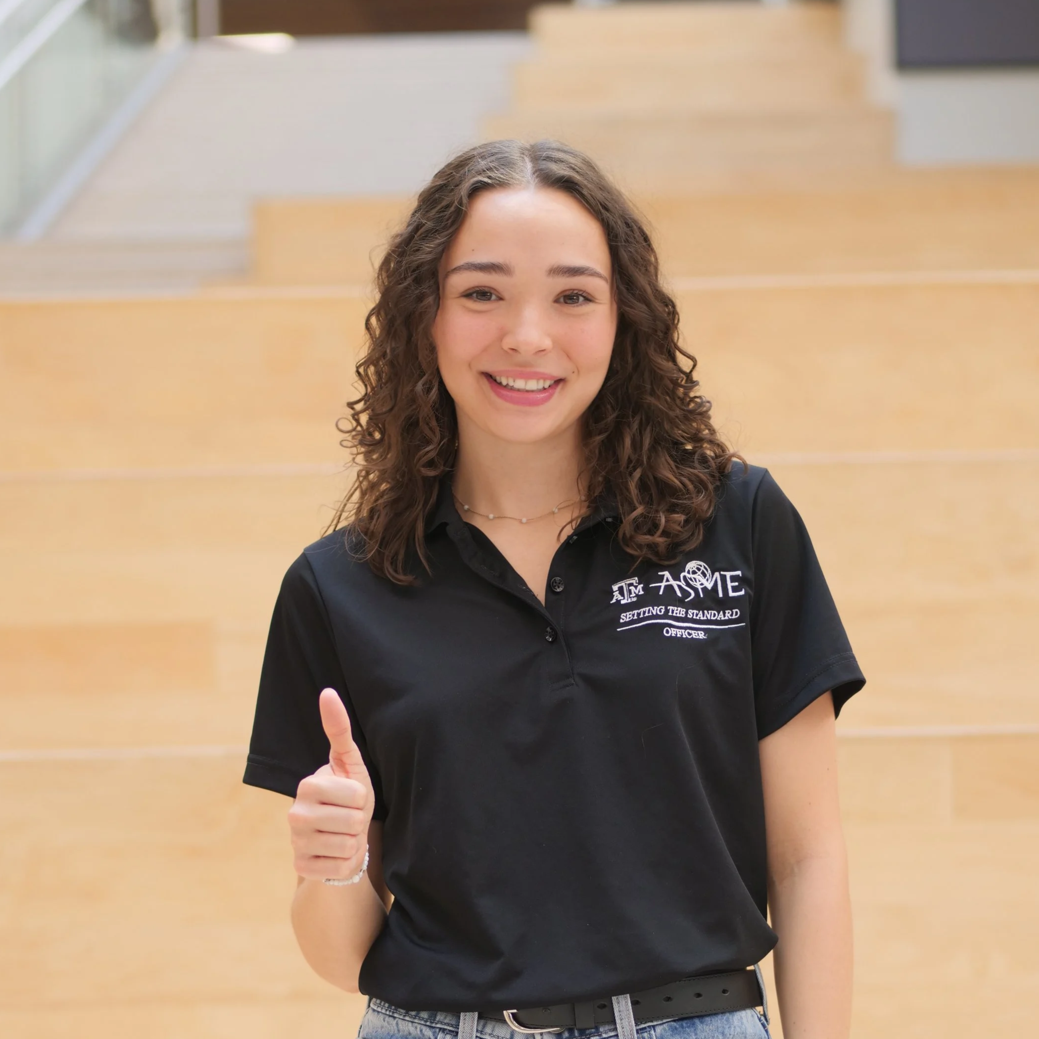 A young woman with curly brown hair smiling and giving a thumbs-up, wearing a black ASME shirt standing in front of a staircase.