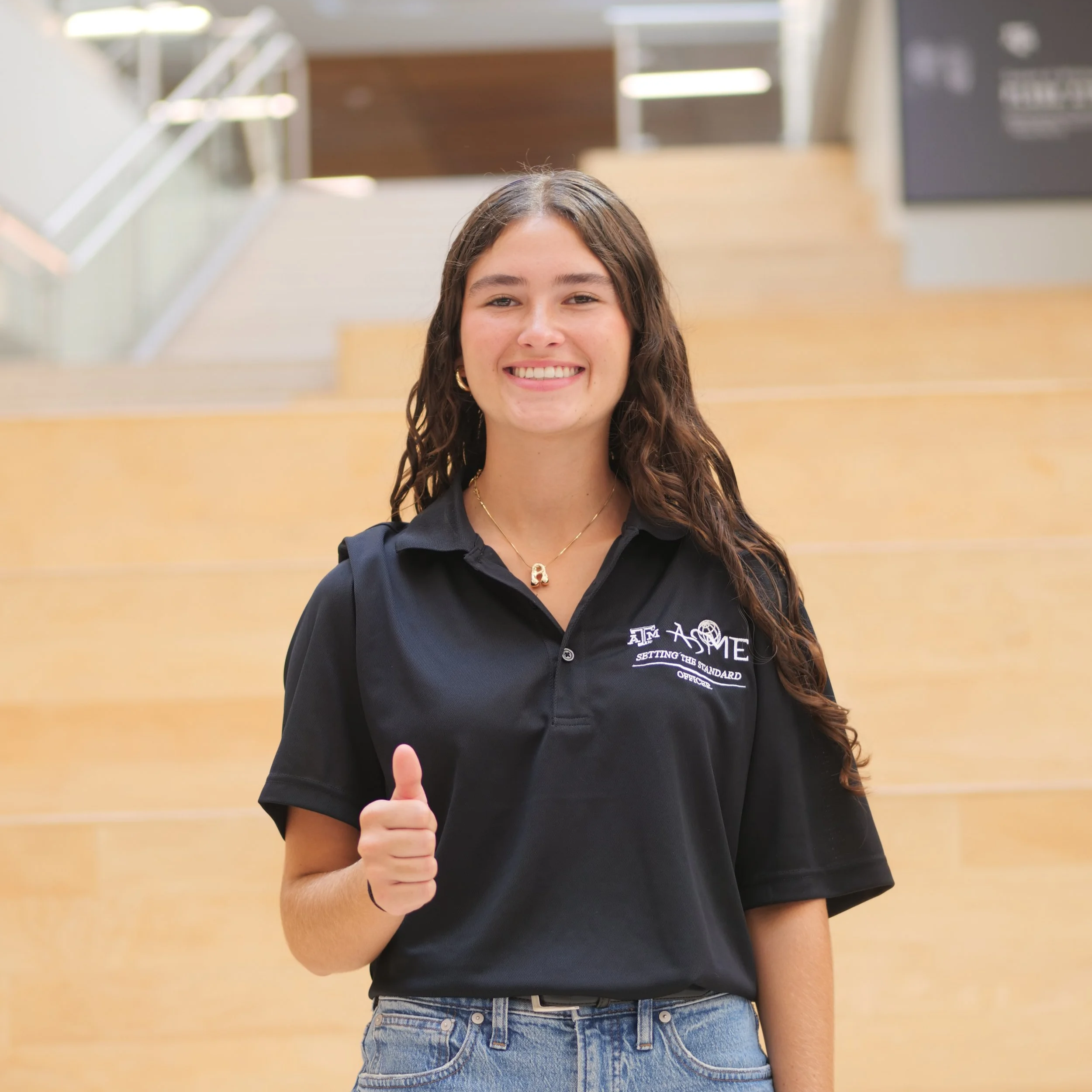 A young woman with long brown hair, wearing a black polo shirt and blue jeans, smiling and giving a thumbs-up in an indoor setting with wooden stairs in the background.