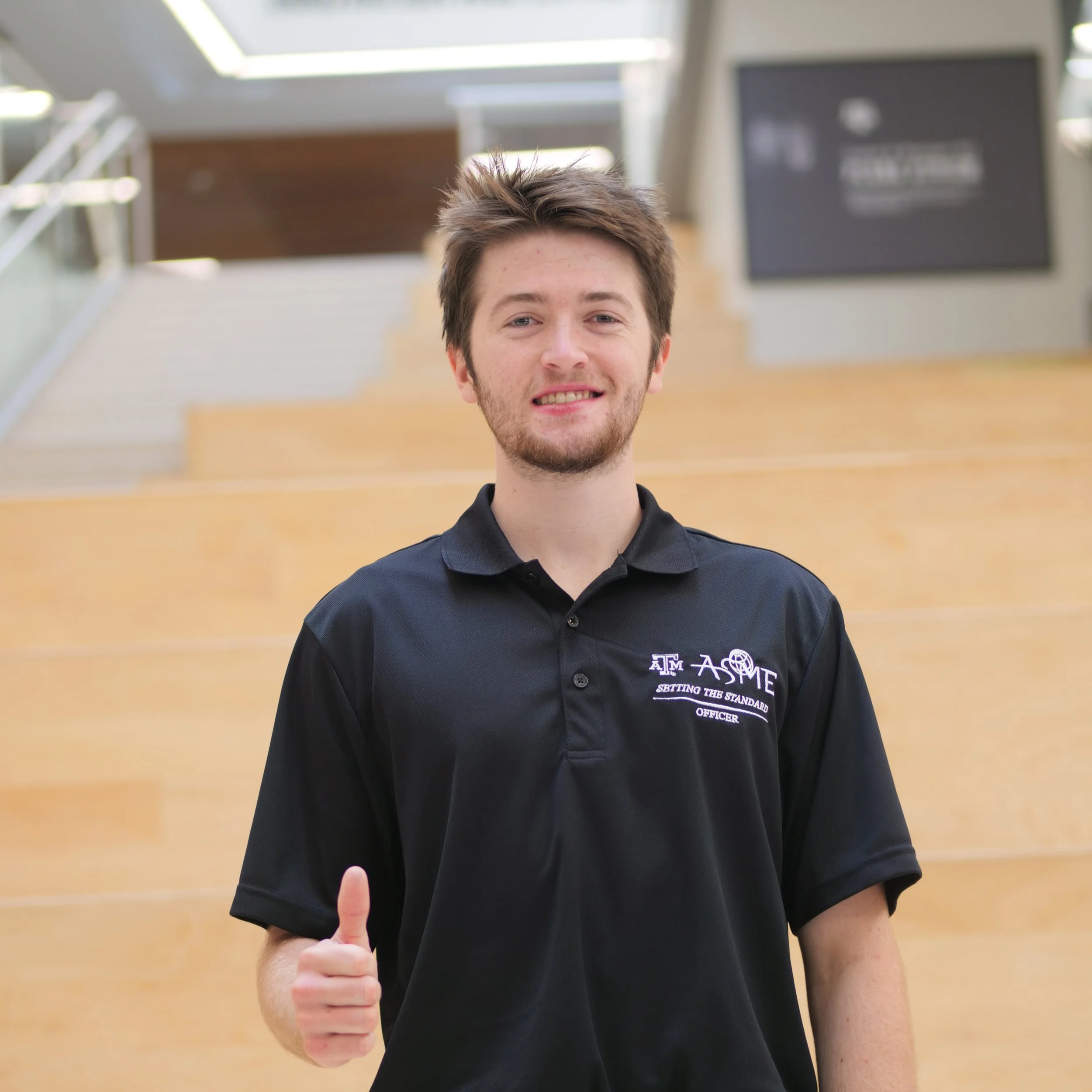 A young man with brown hair and a beard smiling and giving a thumbs-up in a modern indoor space, wearing a black polo shirt with university logos and text.