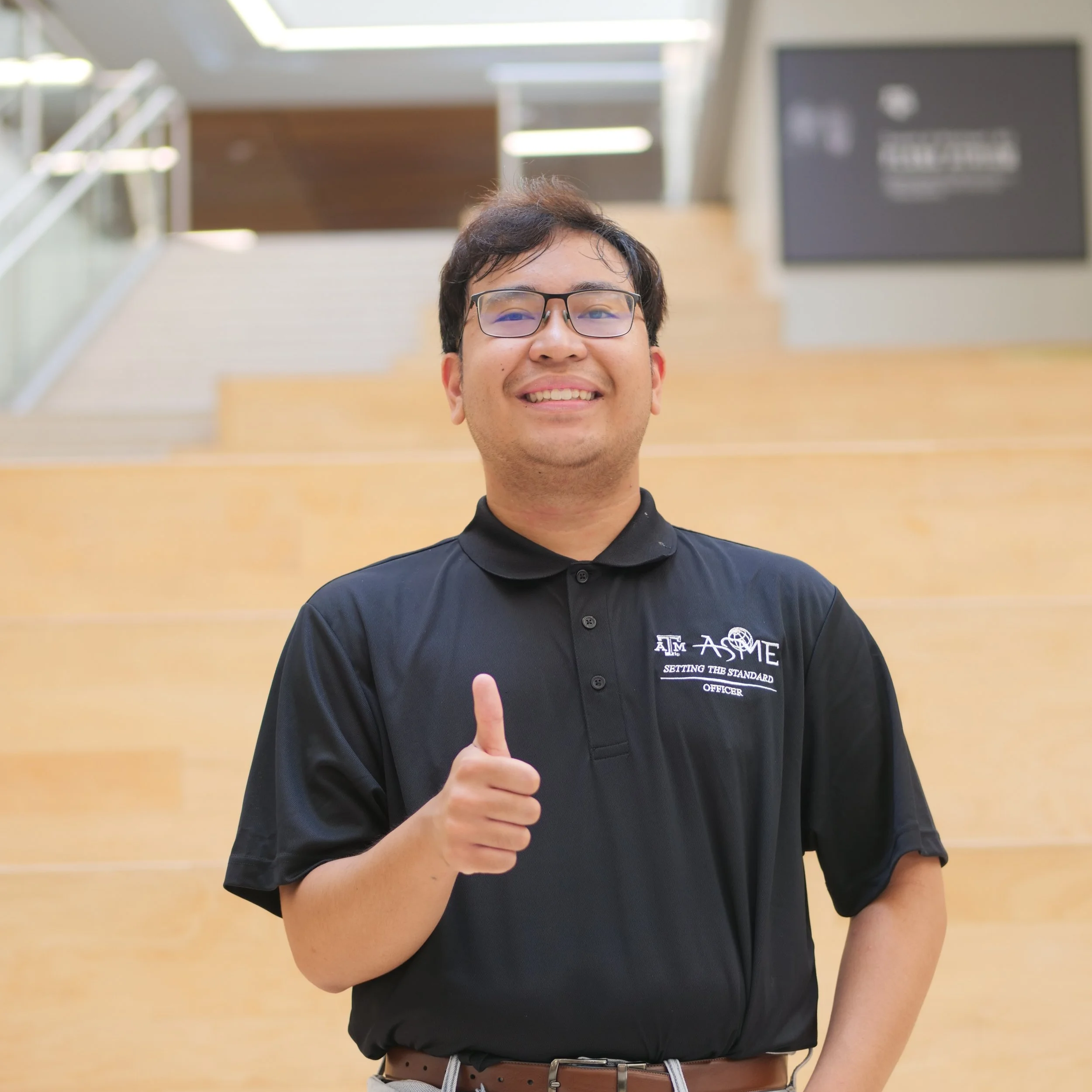 A smiling man wearing glasses and a black polo shirt giving a thumbs-up in an indoor setting with wooden stairs and a blurred background.