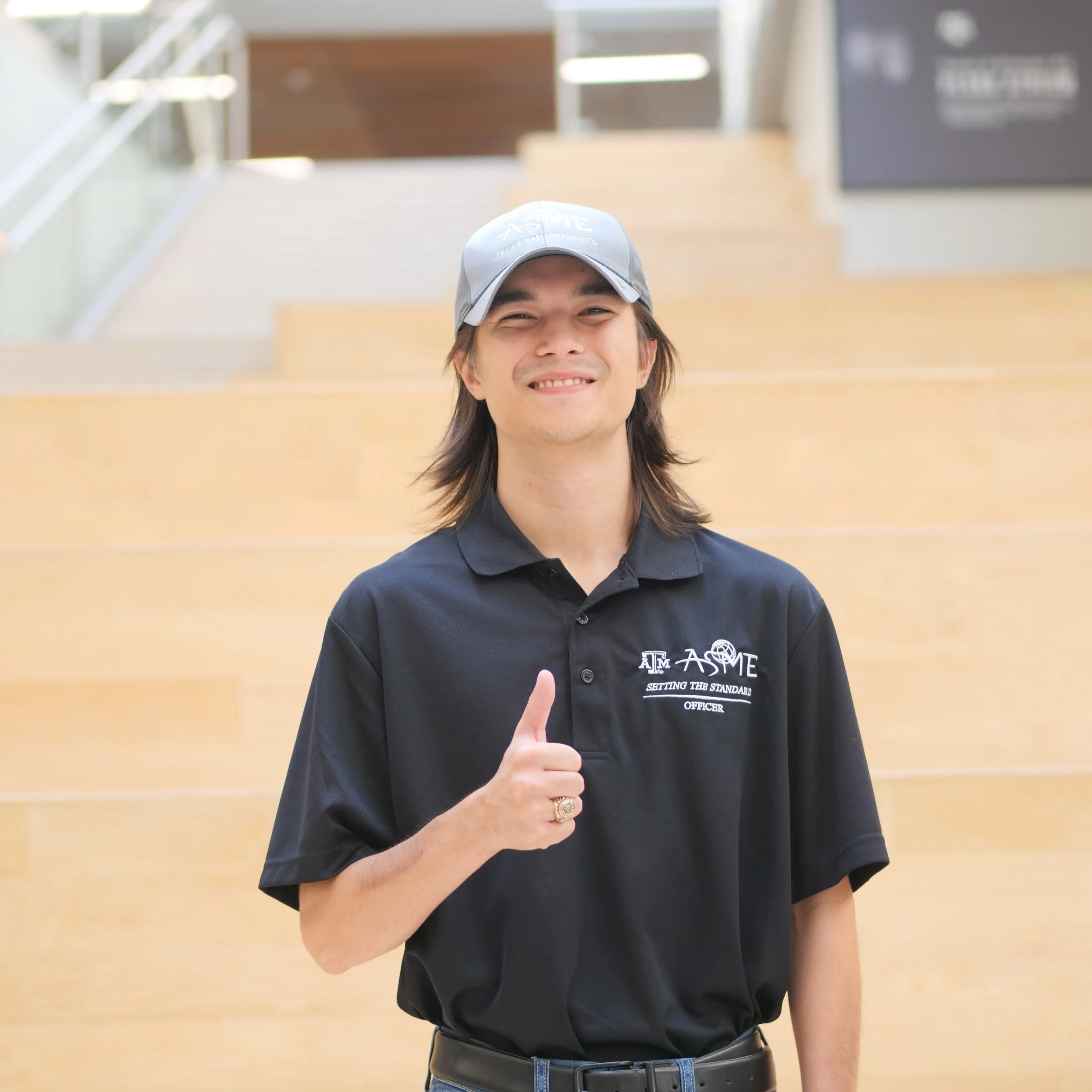 A smiling man in a black polo shirt and a gray cap giving a thumbs-up gesture in an indoor setting with stairs in the background.
