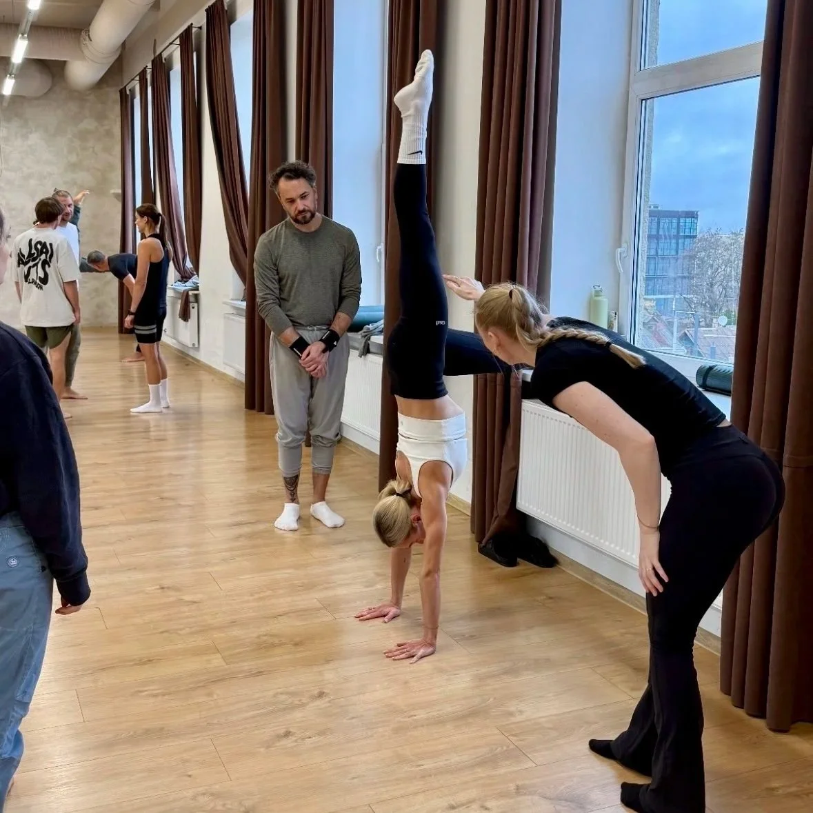 A woman is performing a headstand against a wall while a woman next to her observes. A man stands nearby watching. Other people are engaged in different activities in the background.