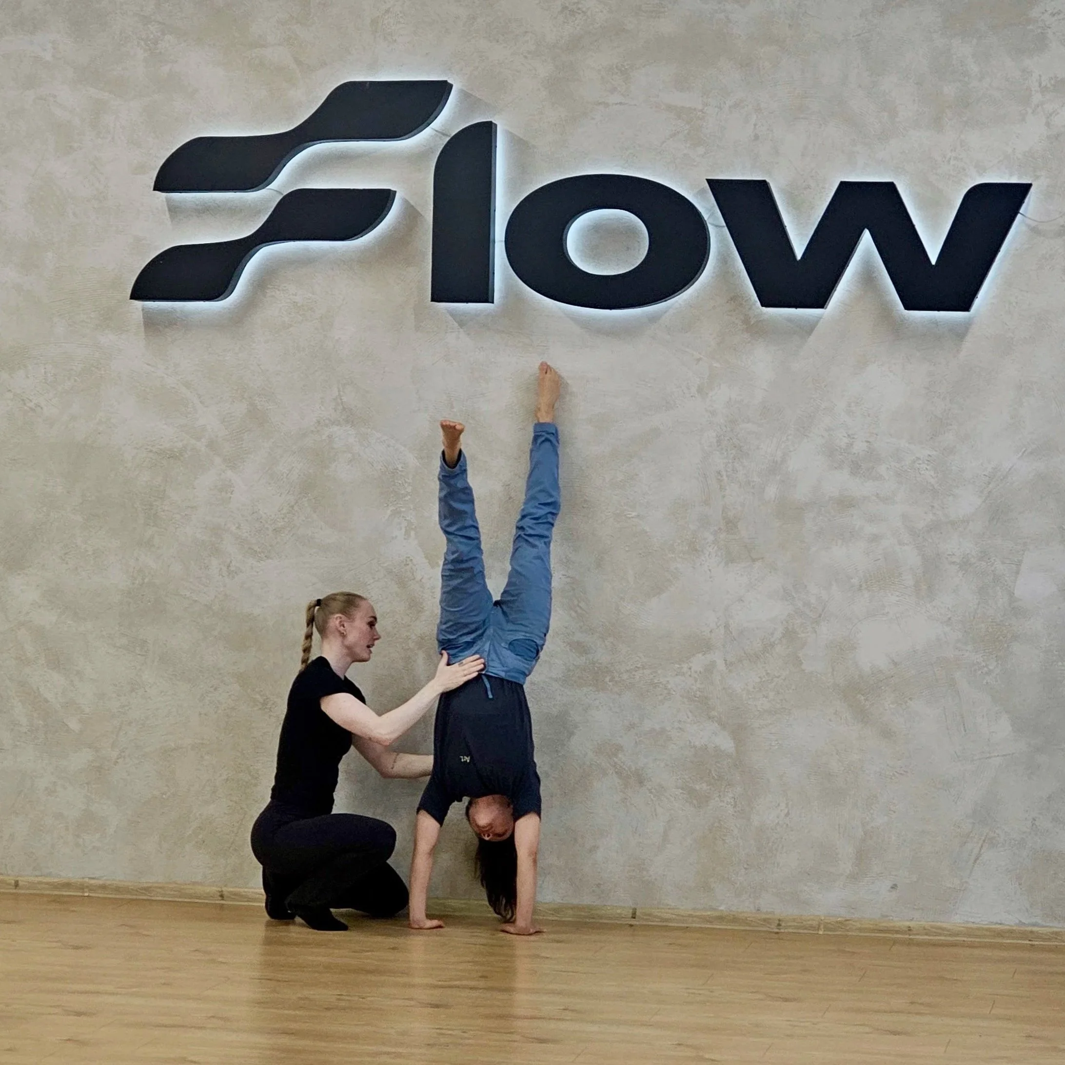 A woman assisting a young girl in performing a handstand in front of a wall with the word 'Flow' and a stylized logo.