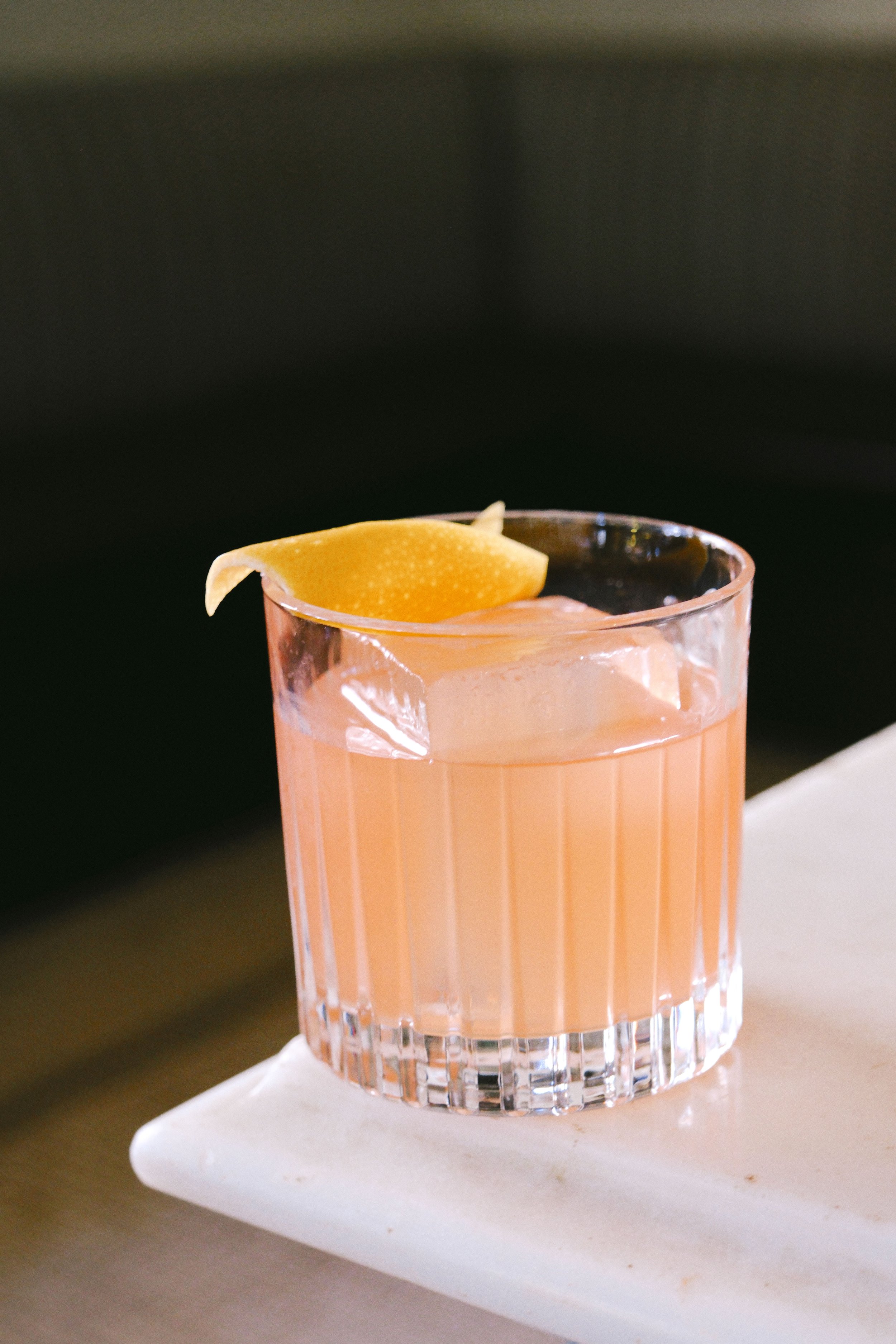 A close-up of a pink colored cocktail with a slice of lemon on top, served in a short, ridged glass placed on a white rectangular plate.