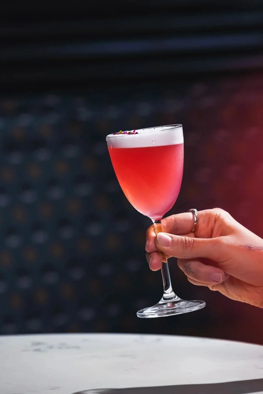 A person's hand holding a pink cocktail in a stemmed glass with a foamy top and small garnish, against a dark background during the best Happy Hour in Yaletown Vancouver at Capo and The Spritz.