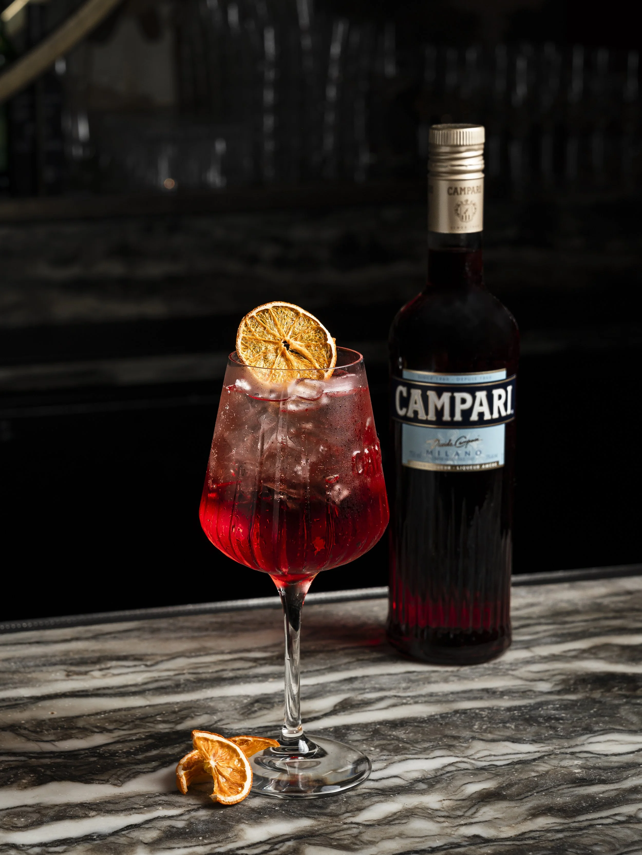 A cocktail in a tall glass with ice and a dried orange slice garnish, set on a marble surface with a bottle of Campari in the background during the best Happy Hour in Yaletown Vancouver at Capo and The Spritz.