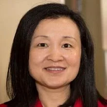 Portrait of a woman with straight black hair and a red top, smiling, set against a neutral indoor background.