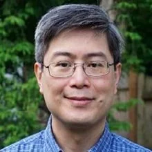 Portrait of a man with glasses and gray hair, wearing a blue checkered shirt, standing outdoors with greenery in the background.