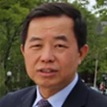 A man in a suit standing outdoors with trees in the background.