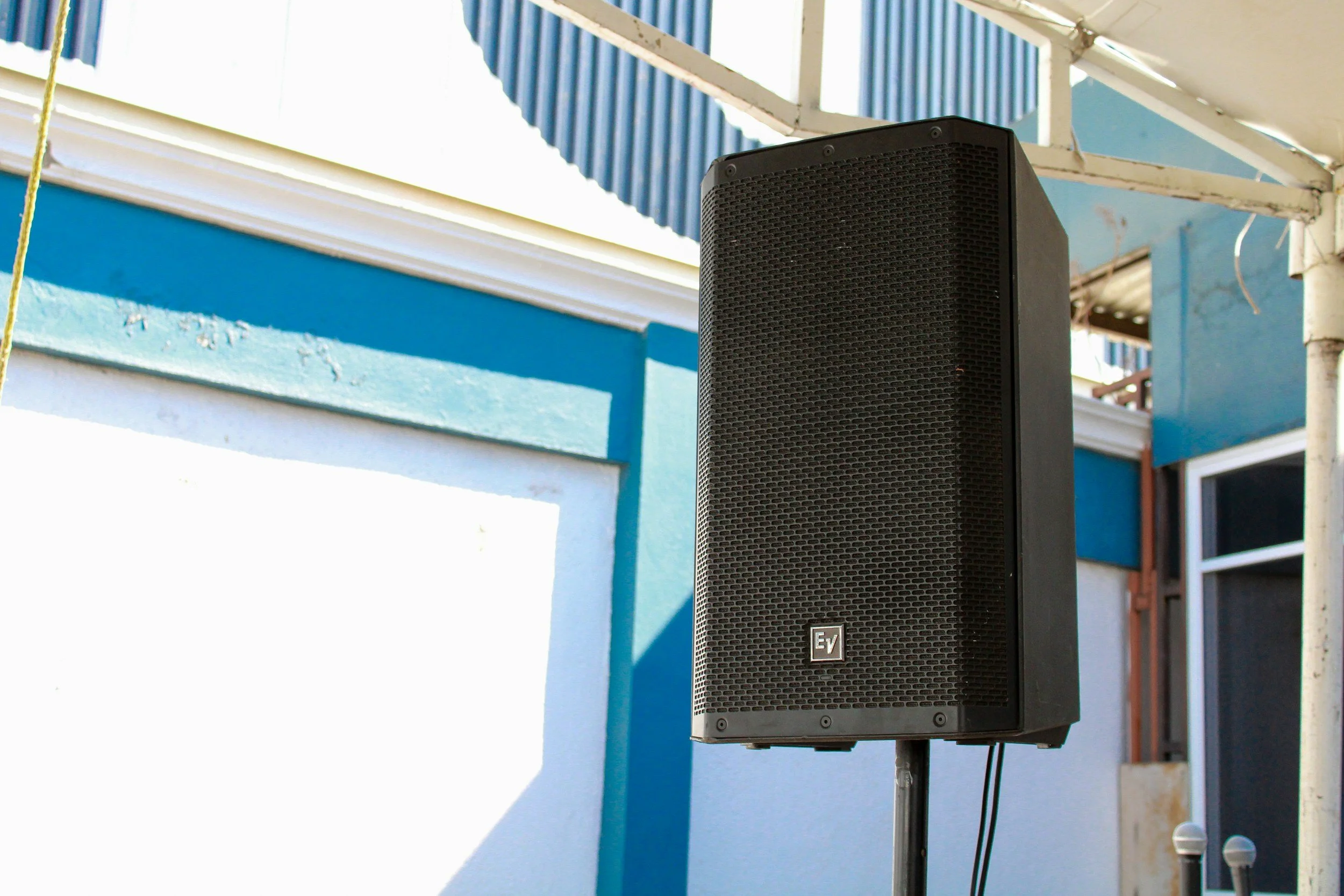 A black EV speaker mounted outside a light blue building with a white trim, under a white and blue awning.