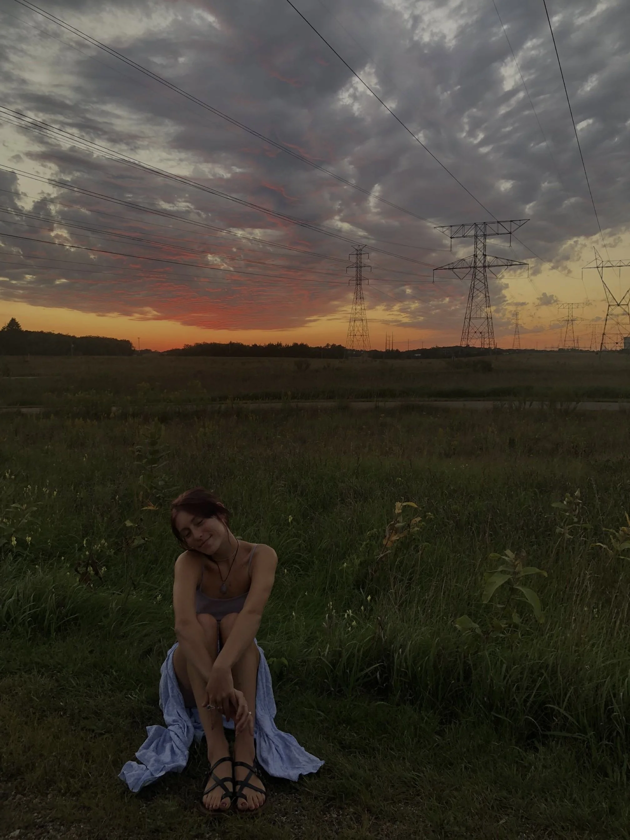 A girl sitting on the grass with a sunset sky and clouds in the background. She is smiling with her eyes closed, wearing a dress and black sandals, with a piece of fabric wrapped around her waist.