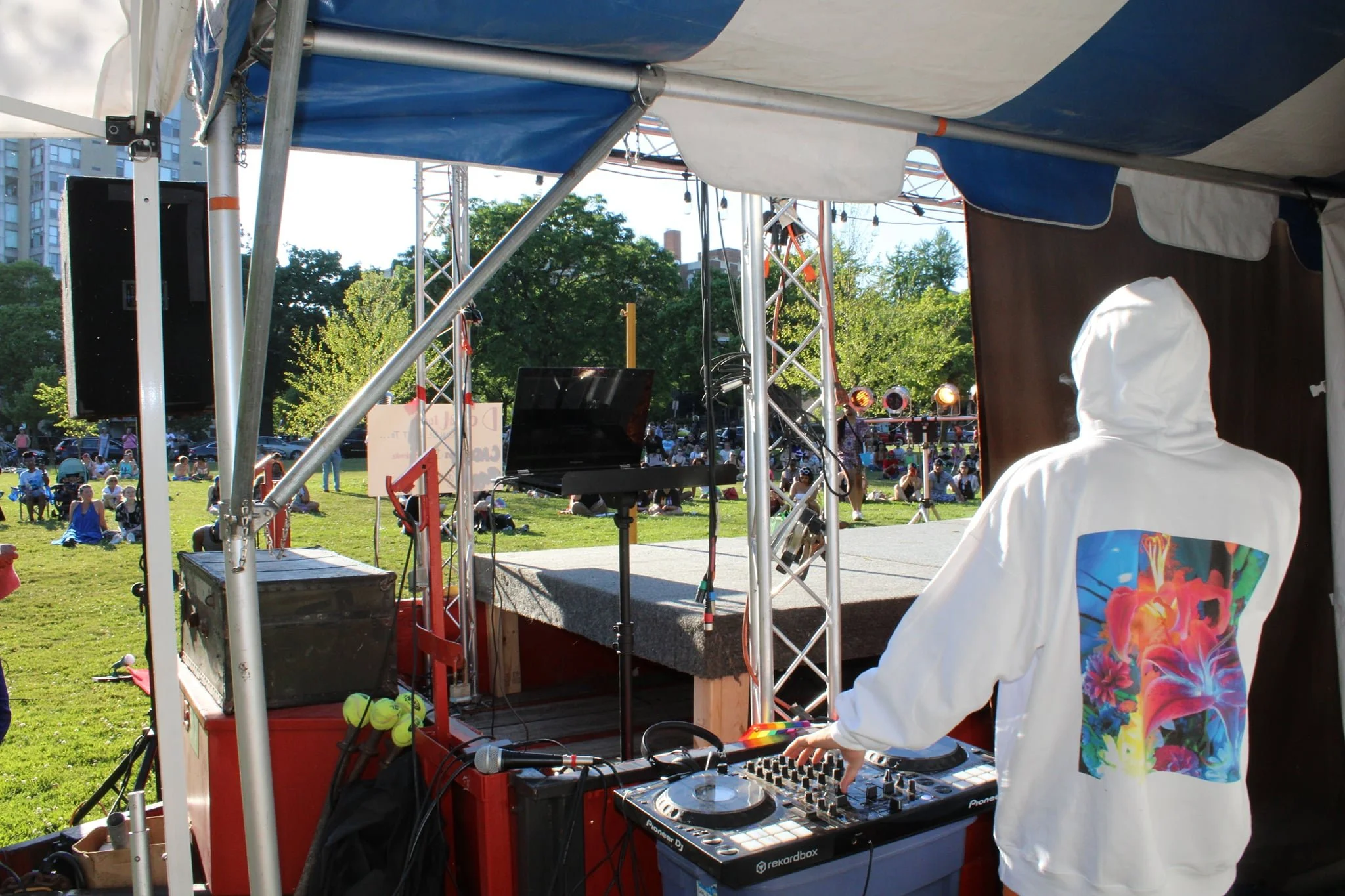 DJ wearing a white hoodie with colorful floral design on the back, operating DJ equipment at an outdoor event in a park with people sitting and walking on the grass, and high-rise buildings in the background.
