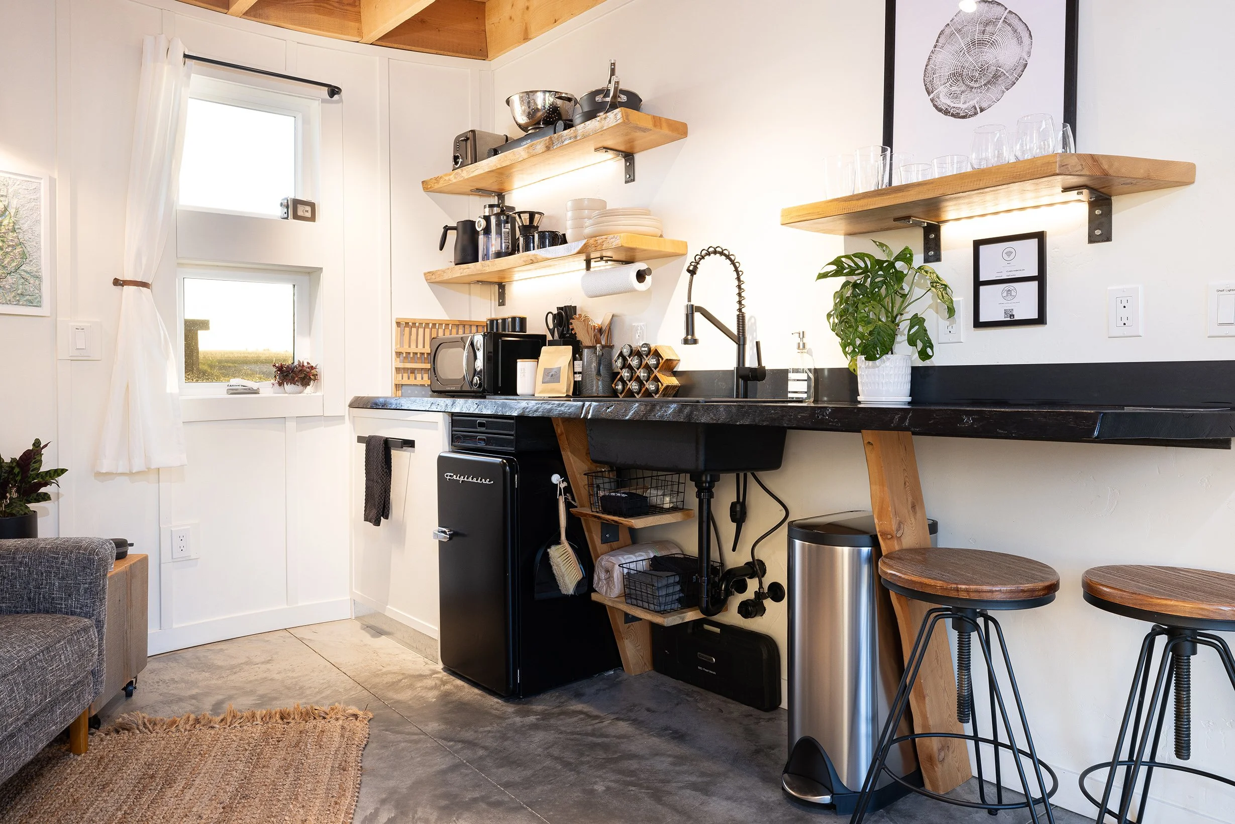Modern kitchen with black natural slab countertop, open wooden shelves, small black refrigerator, white walls, and two wooden bar stools.