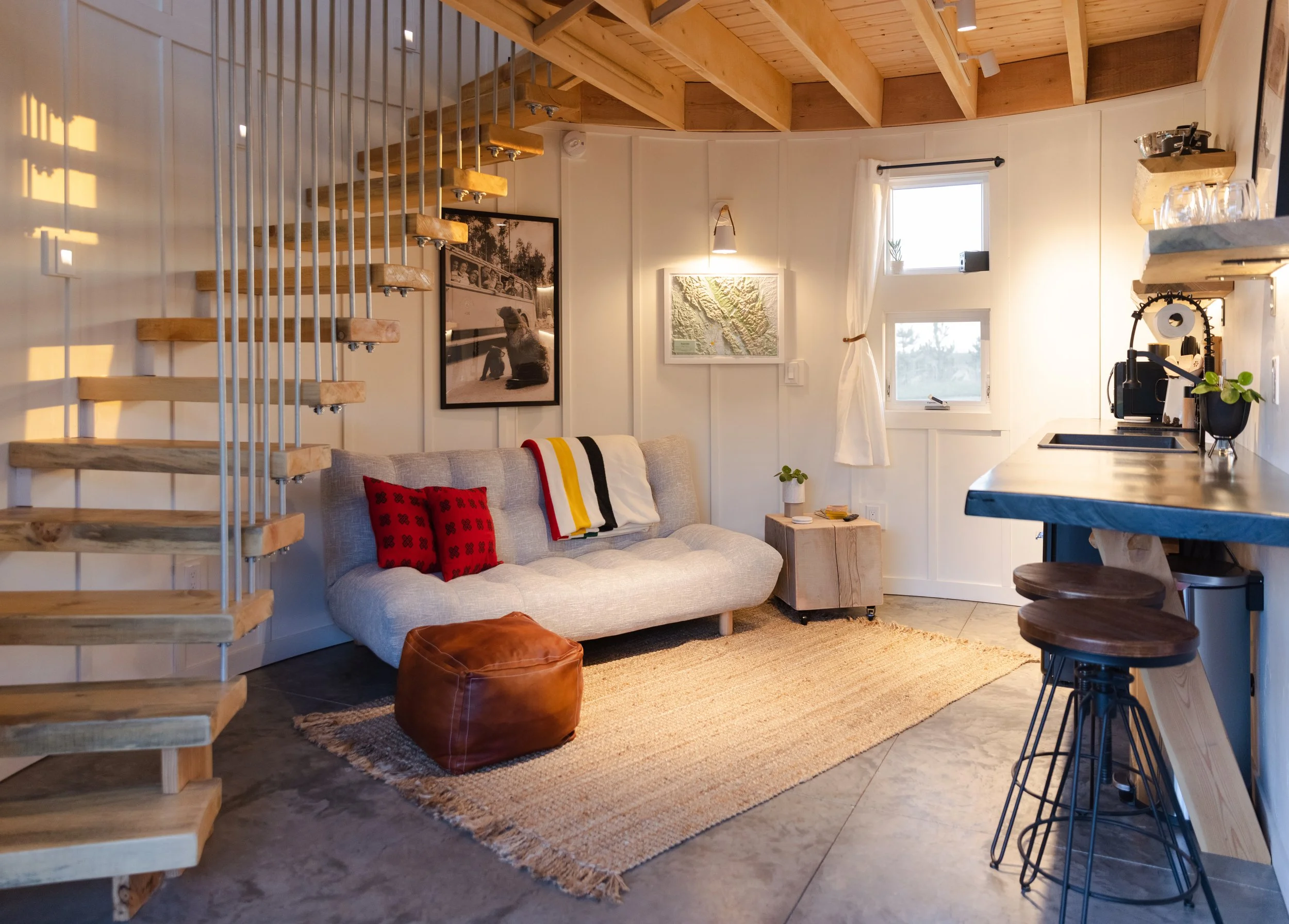 Interior of a cozy living space with a light-colored sofa, red pillows, a leather pouf, a small wooden side table, a rug, a kitchen counter with barstools, and a stairway with wooden steps ascending to a loft area. Natural light comes through small w