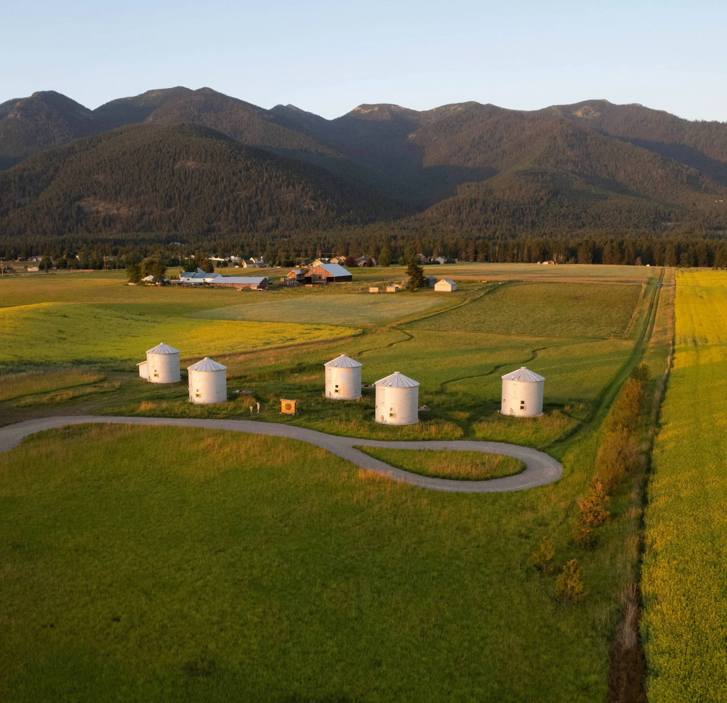 A rural landscape with green fields, six white grain silos, a winding gravel road, farm buildings, and mountains in the background during sunset.