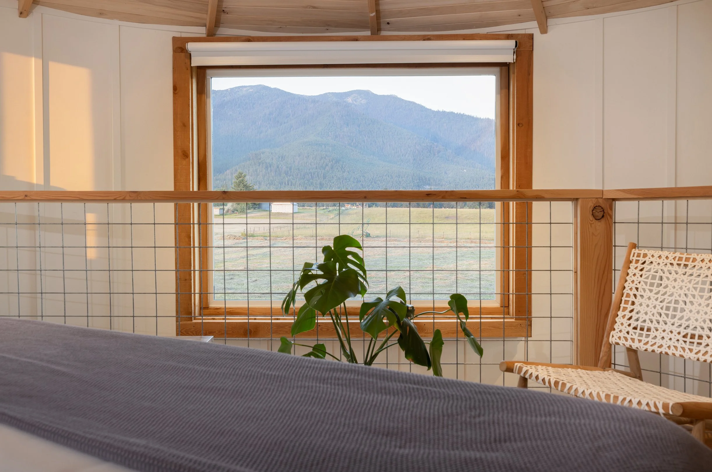 Interior of a room with a large window showing mountains in the background. Inside, there is a wooden chair with a woven seat, a potted plant, and part of a bed with a gray blanket.