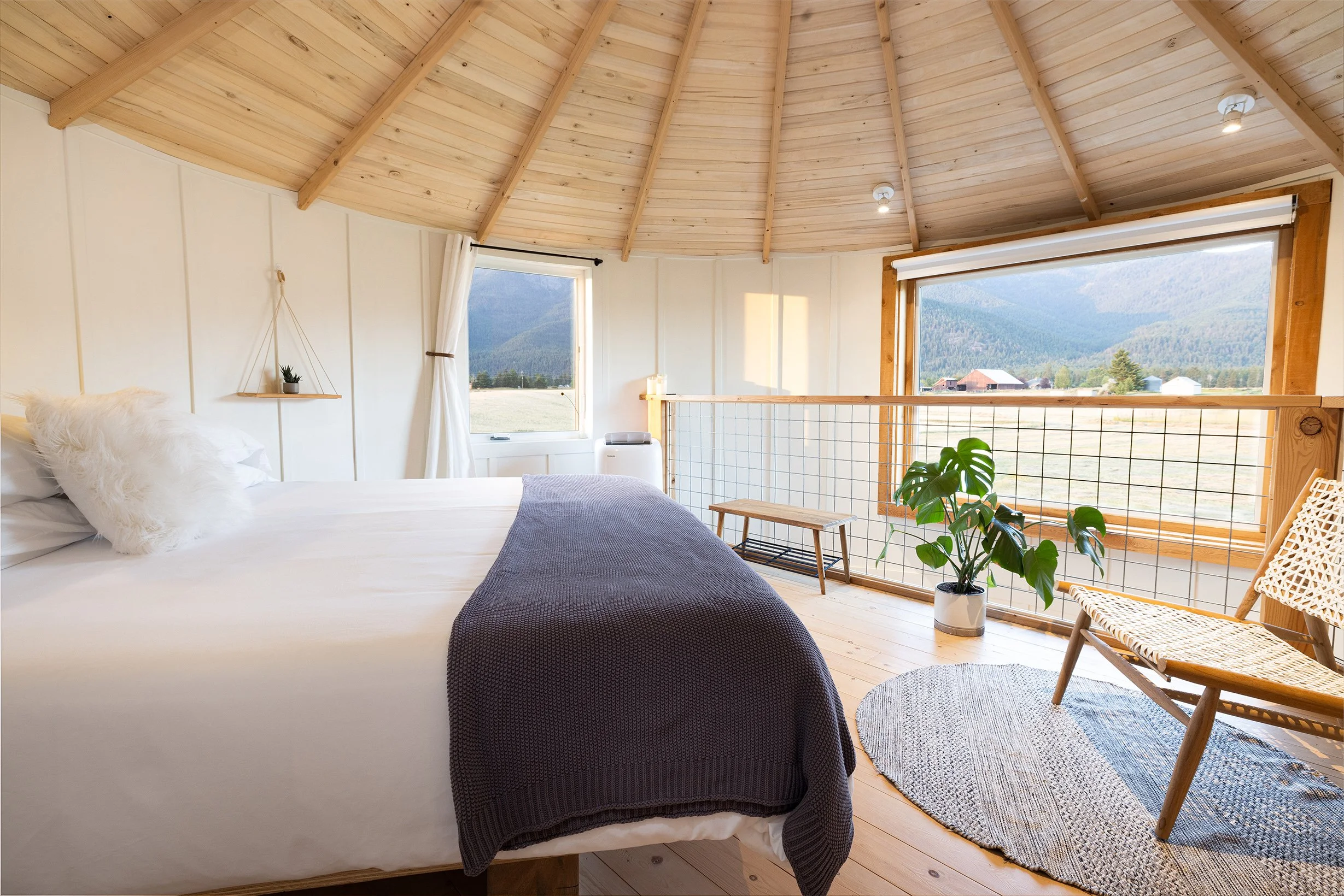 Bedroom with wooden ceiling and large windows showing mountains and fields, white bed with fluffy pillow and gray blanket, potted plant, woven chair, small rug, and bench.