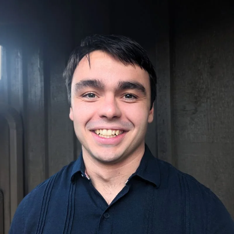 A smiling young man with dark hair wearing a brown blazer and a dark blue shirt against a dark background.
