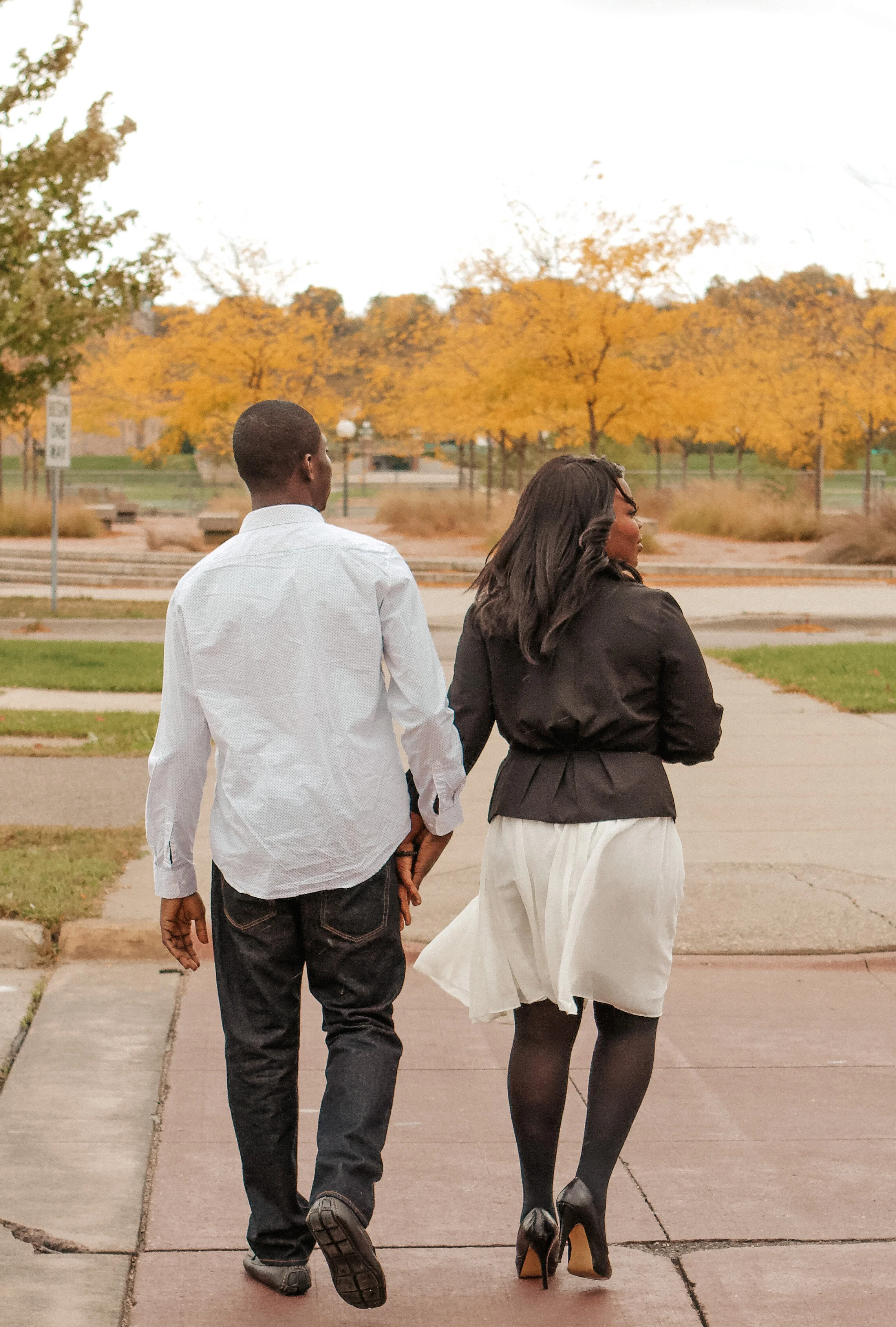 A man and woman walking hand in hand on a sidewalk in a park with orange autumn trees in the background.