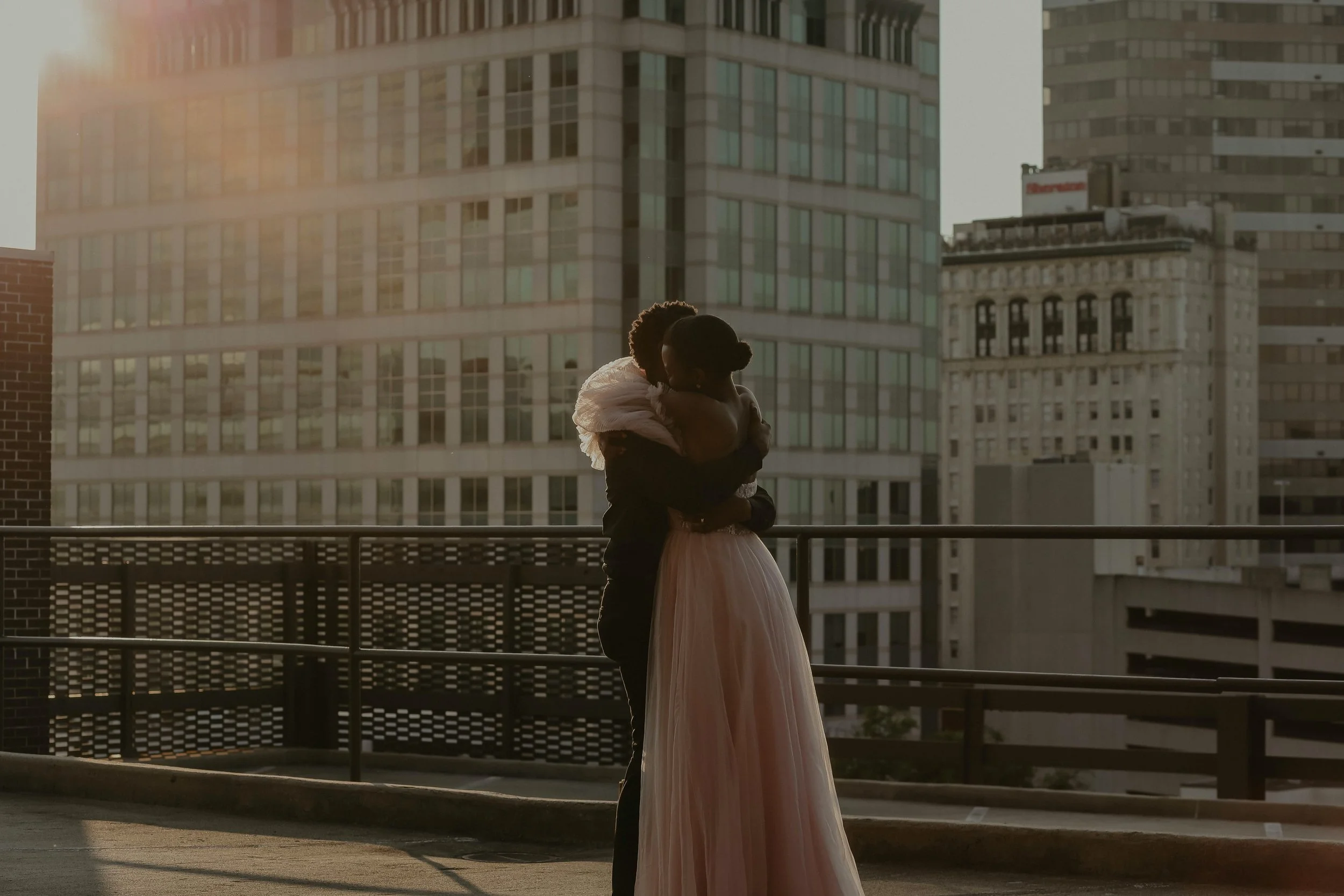 A couple, with the woman wearing a pink dress and the man wrapping his arms around her, standing on a balcony in an urban setting during sunset.