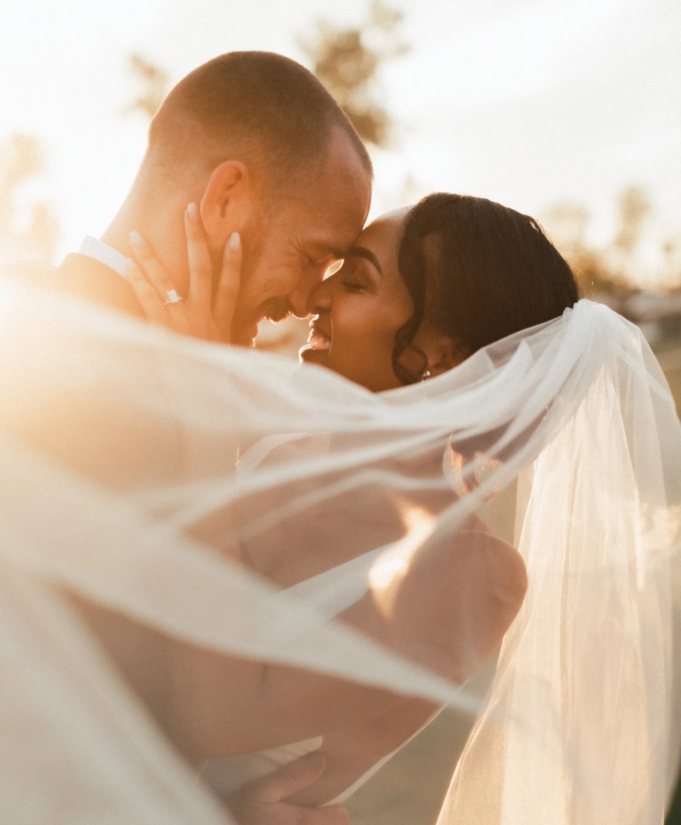 A couple sharing an intimate moment, touching foreheads and smiling, with sunset lighting in the background.